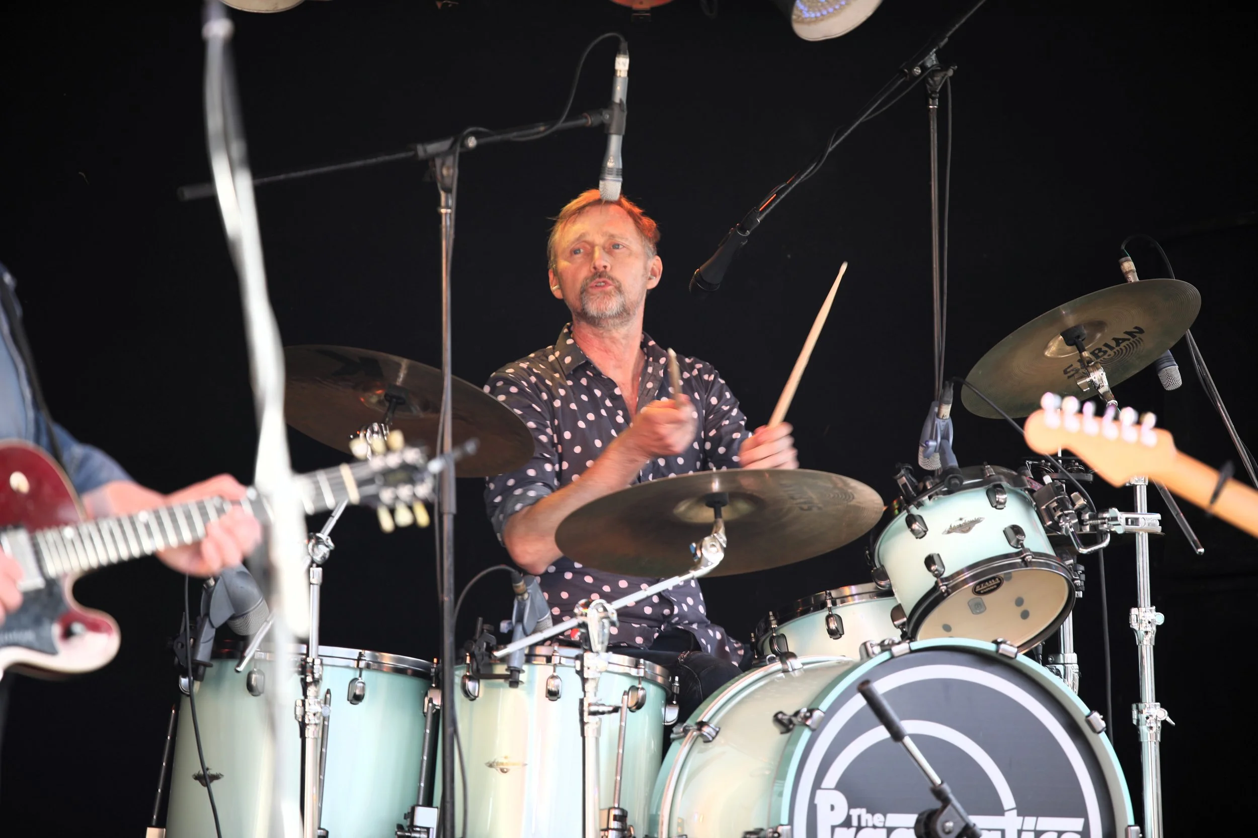 A drummer playing a white drum set during a live performance, with microphones set up overhead and other musical instruments visible.