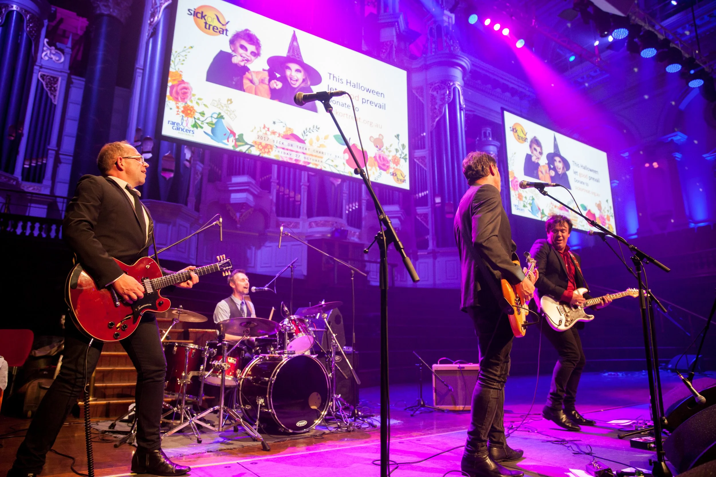 A band performing on stage with purple and blue lighting, with two large screens in the background displaying a Halloween-themed graphic and text about a Halloween event.