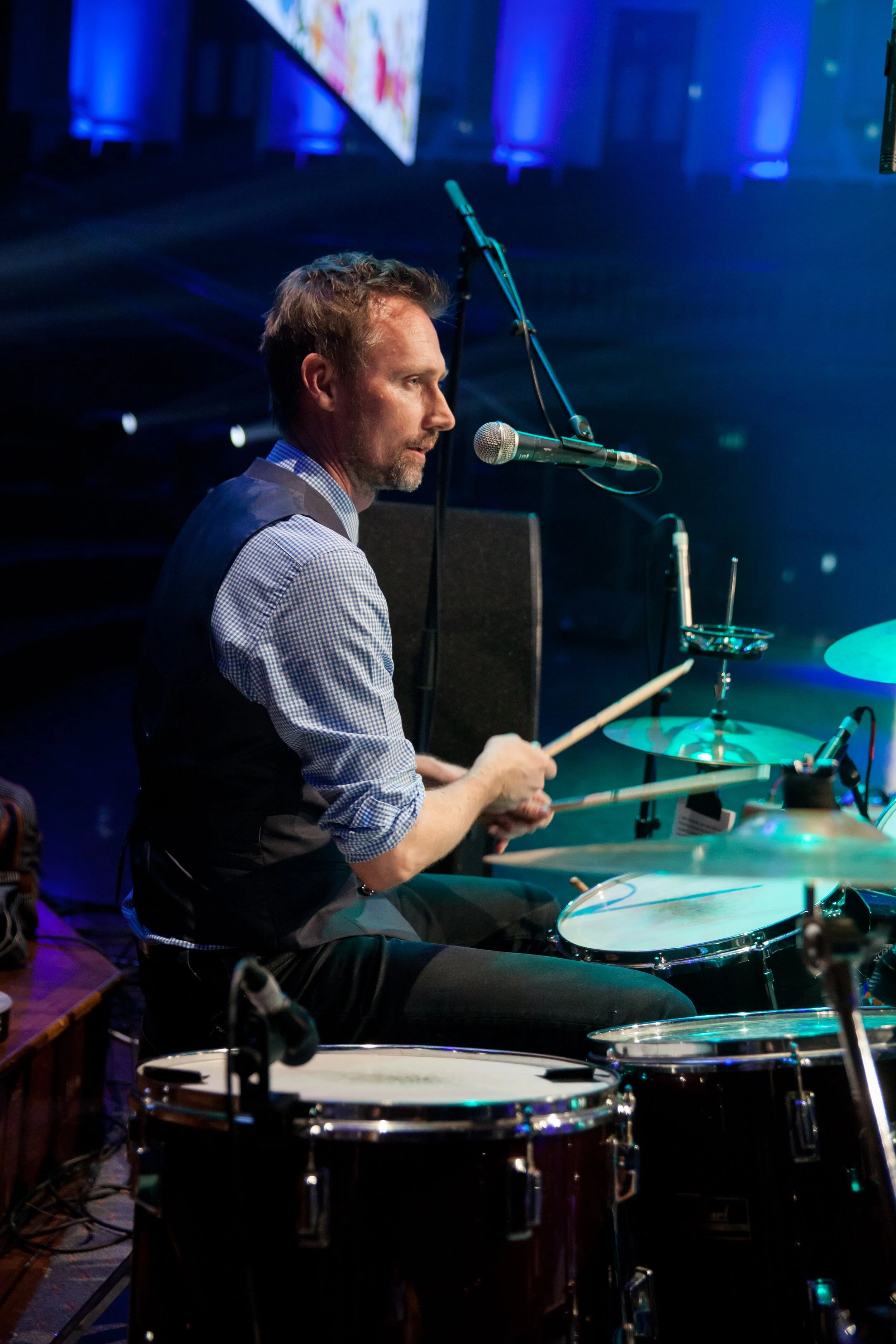 A man playing drums on stage with microphones and drums, illuminated by colorful stage lights at a concert.