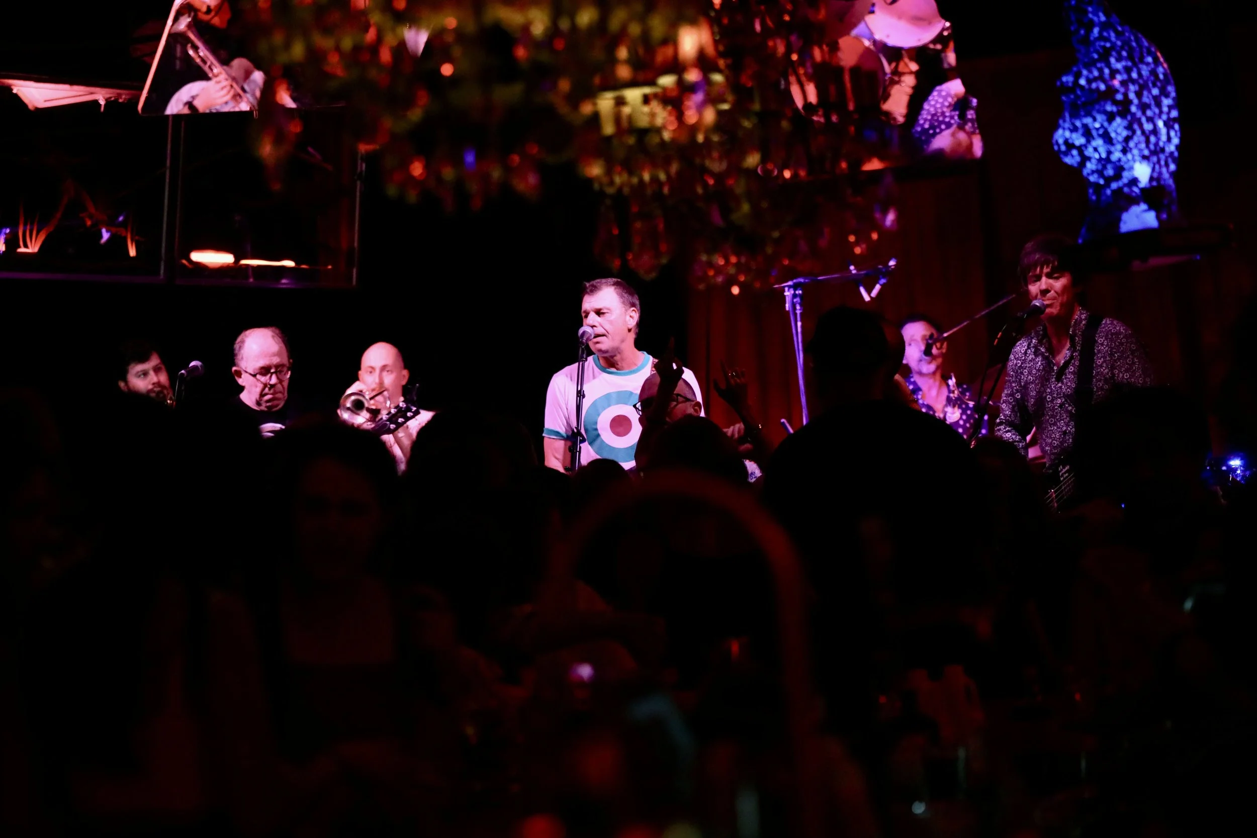 A live music performance at a dimly lit venue with band members playing instruments on stage. Audience members are visible in the foreground.