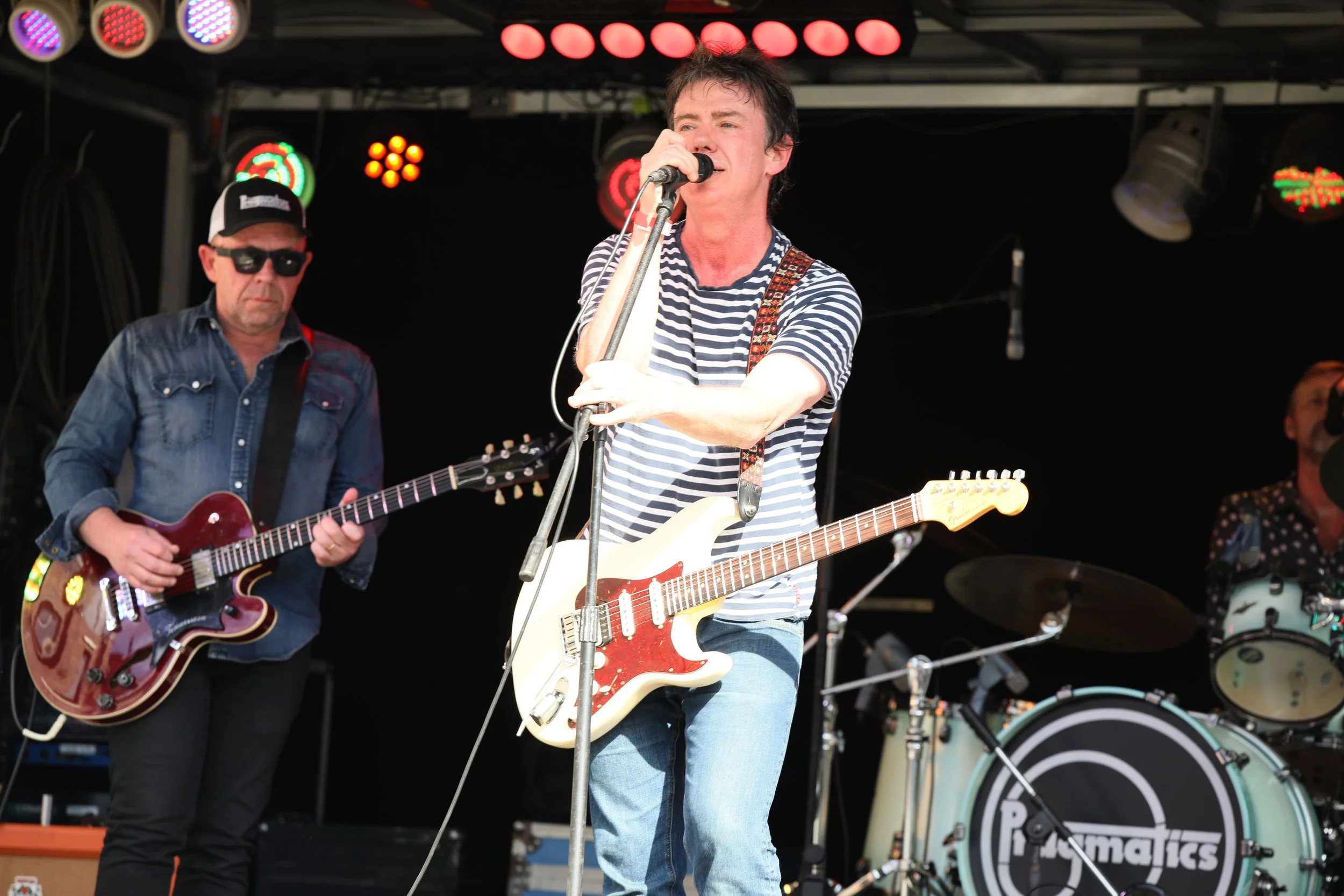 A band performing on stage with guitarists and a drummer, stage lights overhead, one guitarist holding a white electric guitar and singing into a microphone.