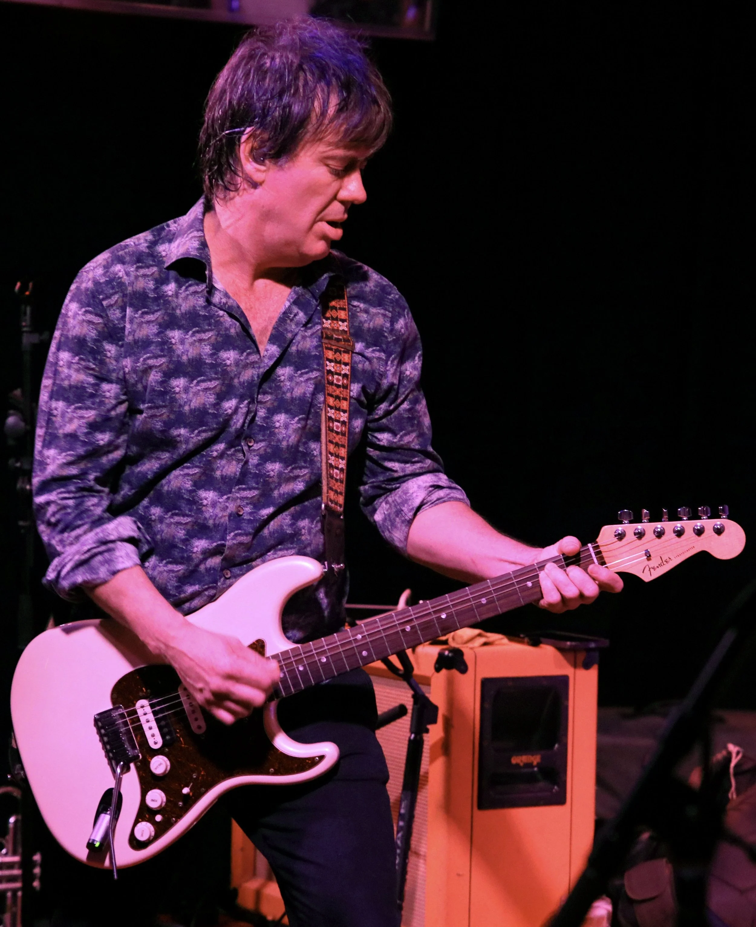 A man playing an electric guitar on stage with purple lighting.