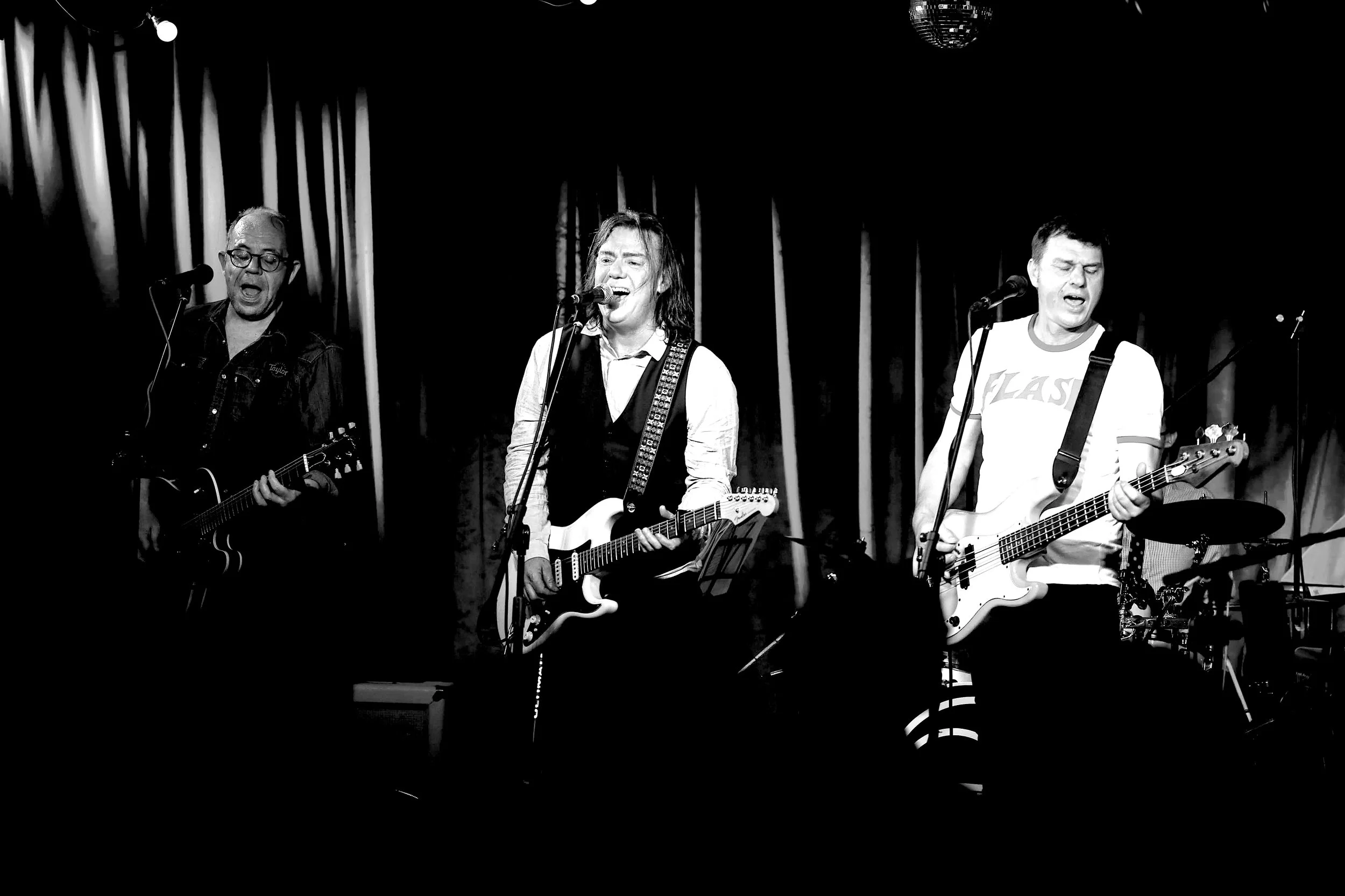 A black and white photo of three musicians on a stage playing guitars and singing with a dark curtain backdrop.