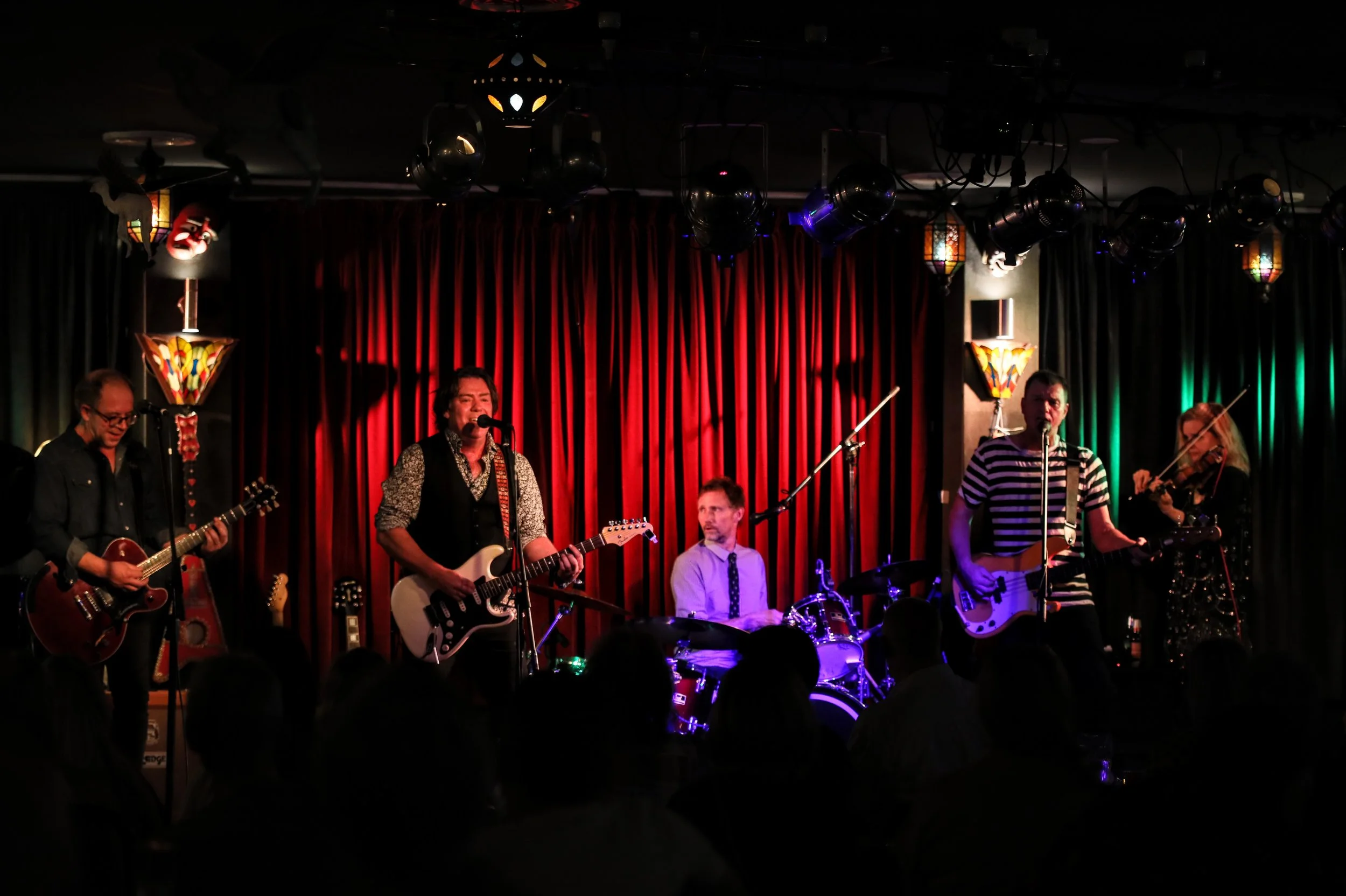 A band performing on stage with red curtains and colorful lamps, featuring five musicians playing guitar, drums, and violin, with a dark audience in front.