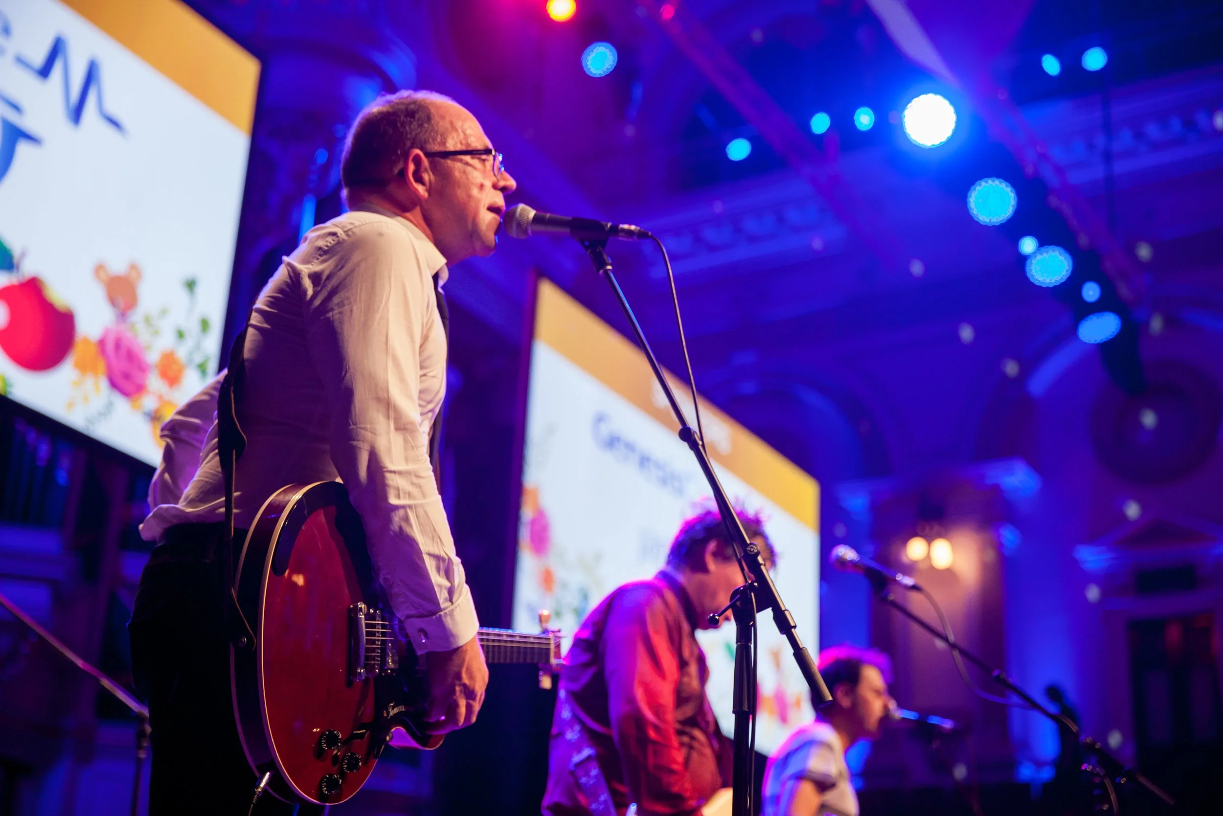 Musicians performing on stage at a concert with colorful lighting and screen in the background.