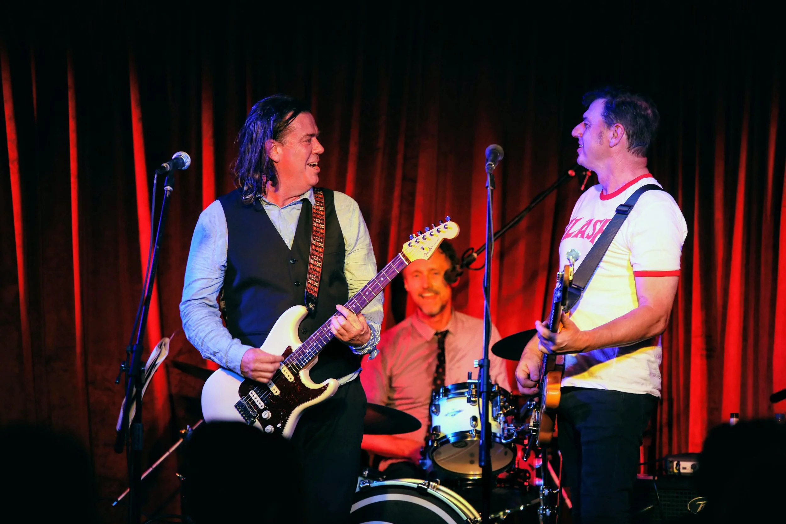 Three male musicians performing on stage with guitars and drums, red curtains in the background, smiling and engaging with each other.