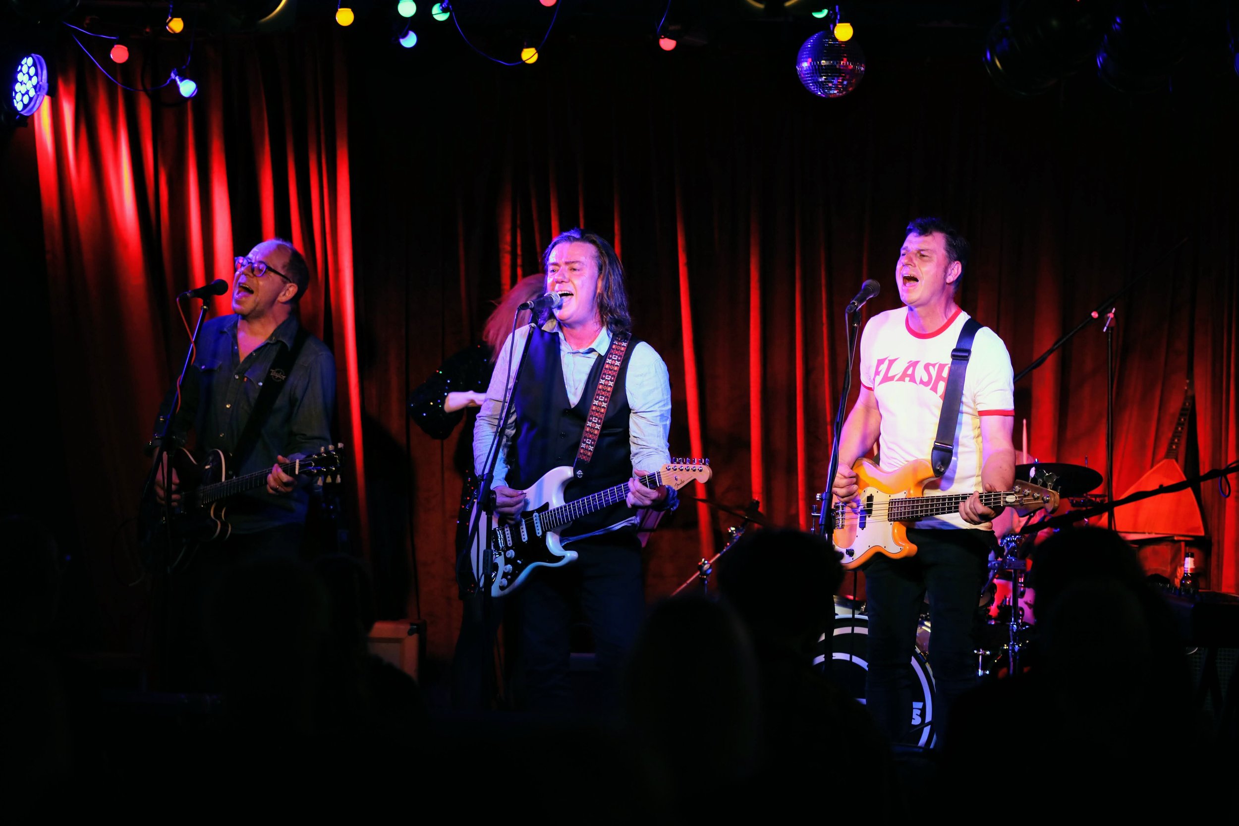 Men playing guitar and singing on stage with red curtain background, colorful stage lights, and audience silhouettes.