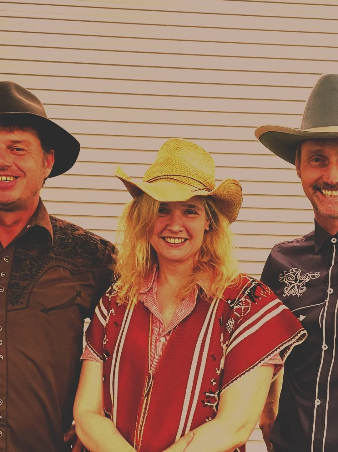 Three people wearing cowboy hats, smiling, standing in front of a white horizontal paneled wall.