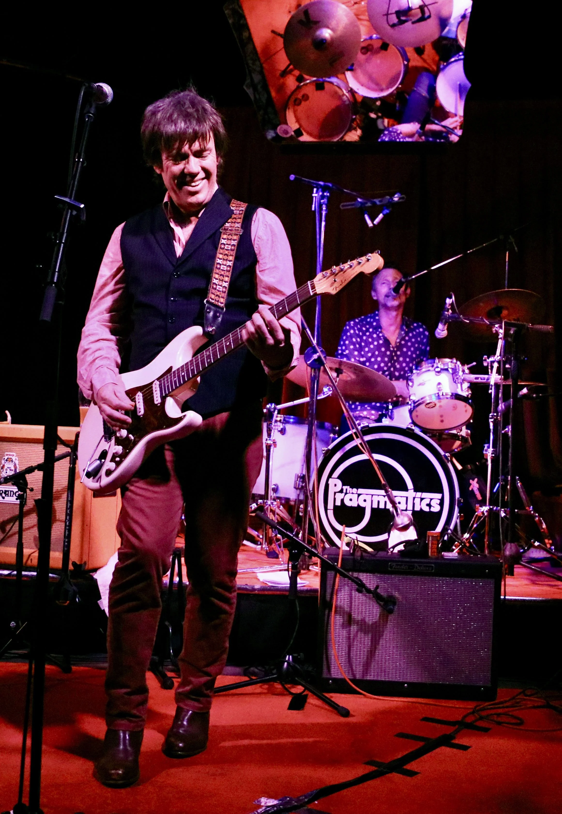 Two musicians performing on stage; one playing an electric guitar and smiling, the other playing drums in the background. The drum kit has the band name 'The Pragmatists' on the bass drum.