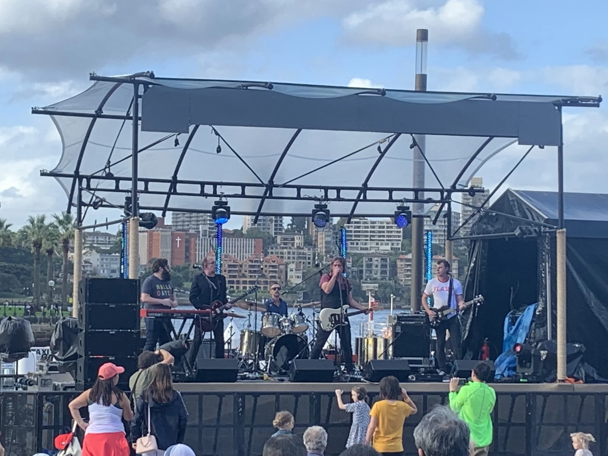 A live outdoor band performs on a stage with five band members, including a drummer, a keyboardist, and three guitarists. Audience members are watching and taking photos. The background features a city skyline with buildings, palm trees, and a cloudy