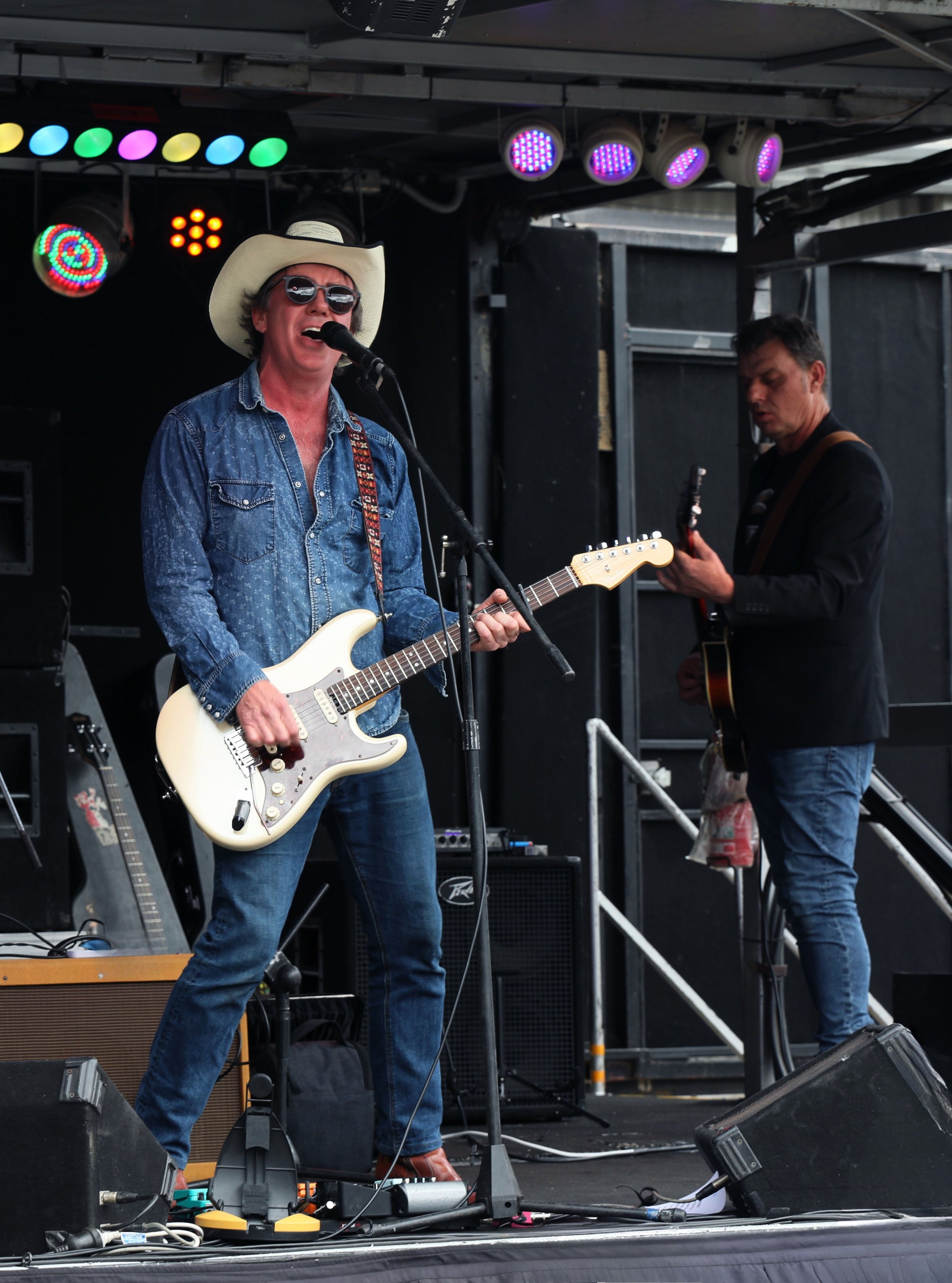 Two musicians performing on a stage: one Hispanic man wearing a cowboy hat, sunglasses, a denim shirt, jeans, and playing an electric guitar, singing into a microphone; the other man, also Hispanic, wearing a black jacket and jeans, playing another g