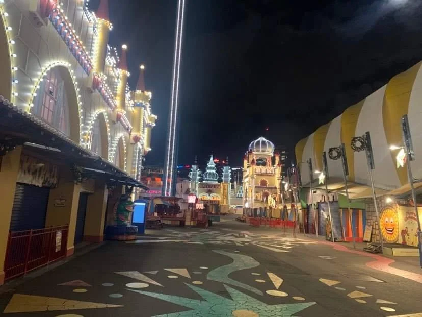 Empty amusement park at night with lit-up buildings and rides, including a castle-like structure and striped tents, no visitors present.