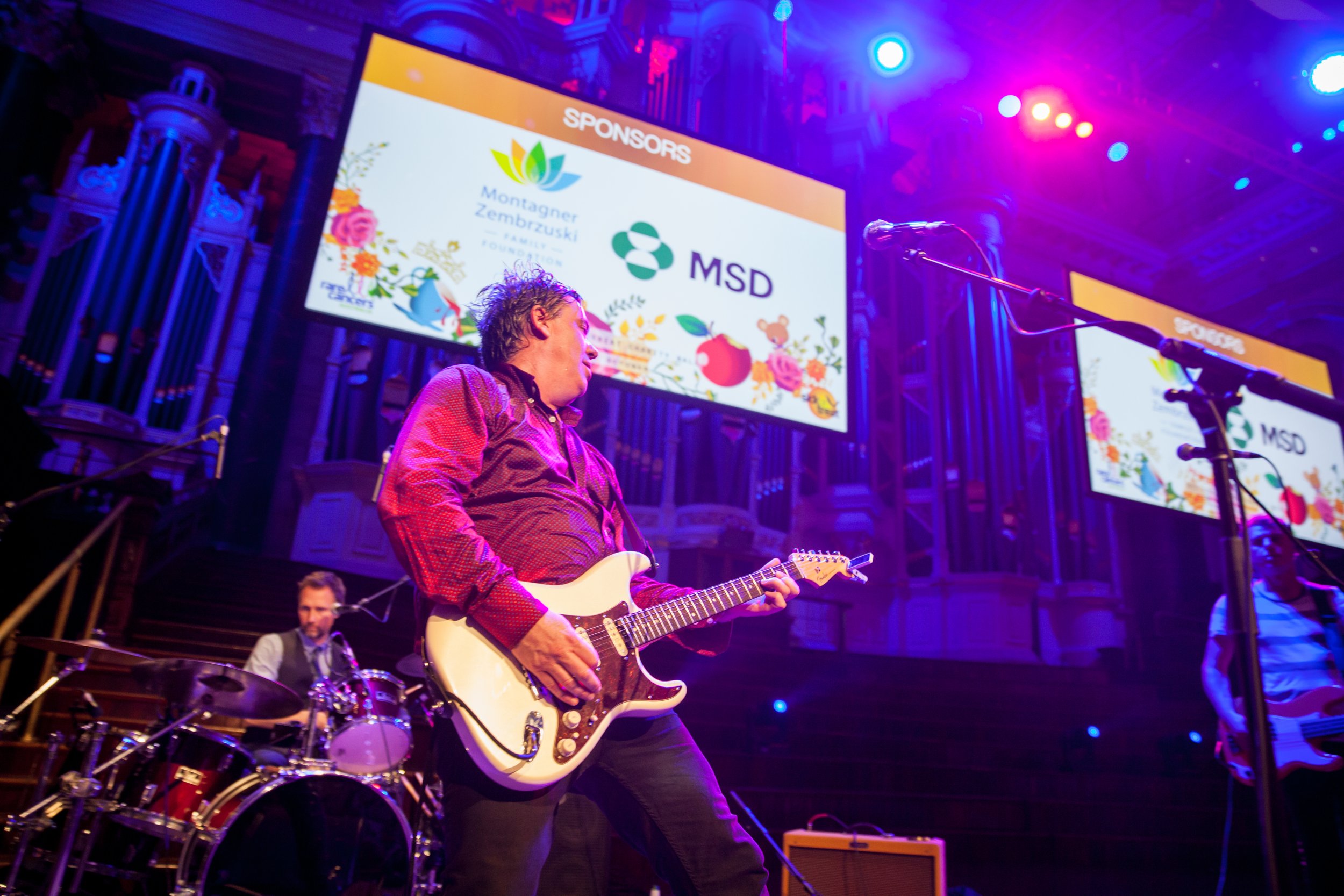Musicians performing on stage with large sponsor screens in the background, featuring colorful floral graphics and logos, including MSD and Montagner Zembruski.