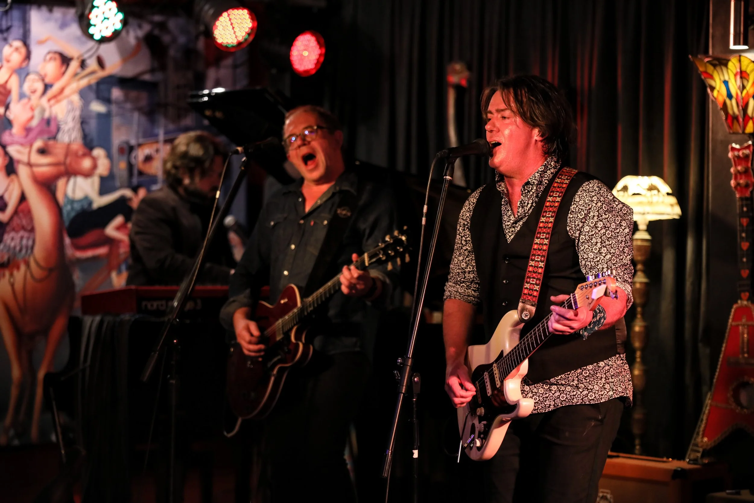 A band of three men performing live on stage with guitars and a keyboard, singing into microphones, with colorful stage lights and vintage decor in the background.