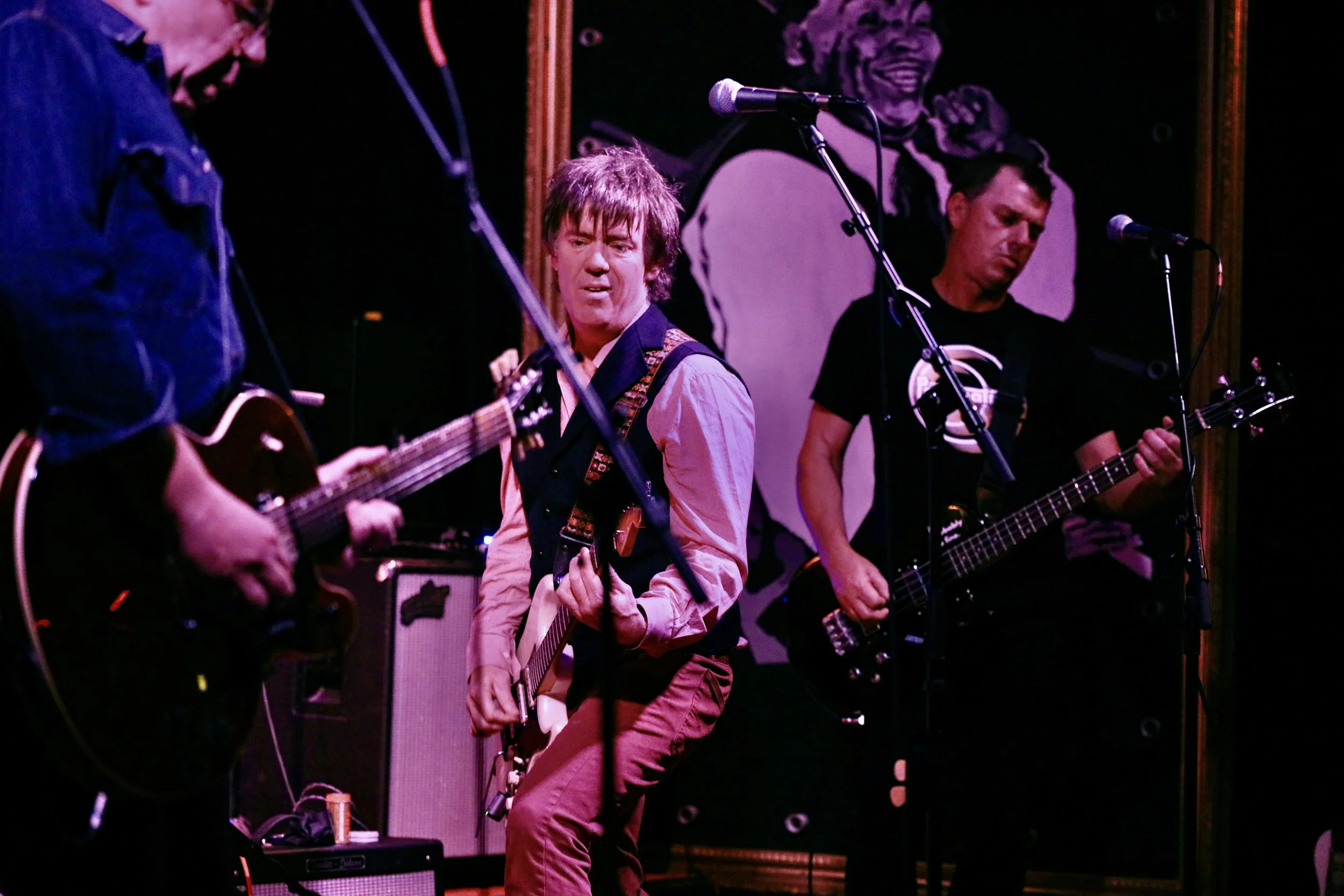Three musicians performing on stage, one playing an acoustic guitar, another playing an electric guitar, and the third playing a bass guitar, with a large black-and-white portrait of a woman in the background.