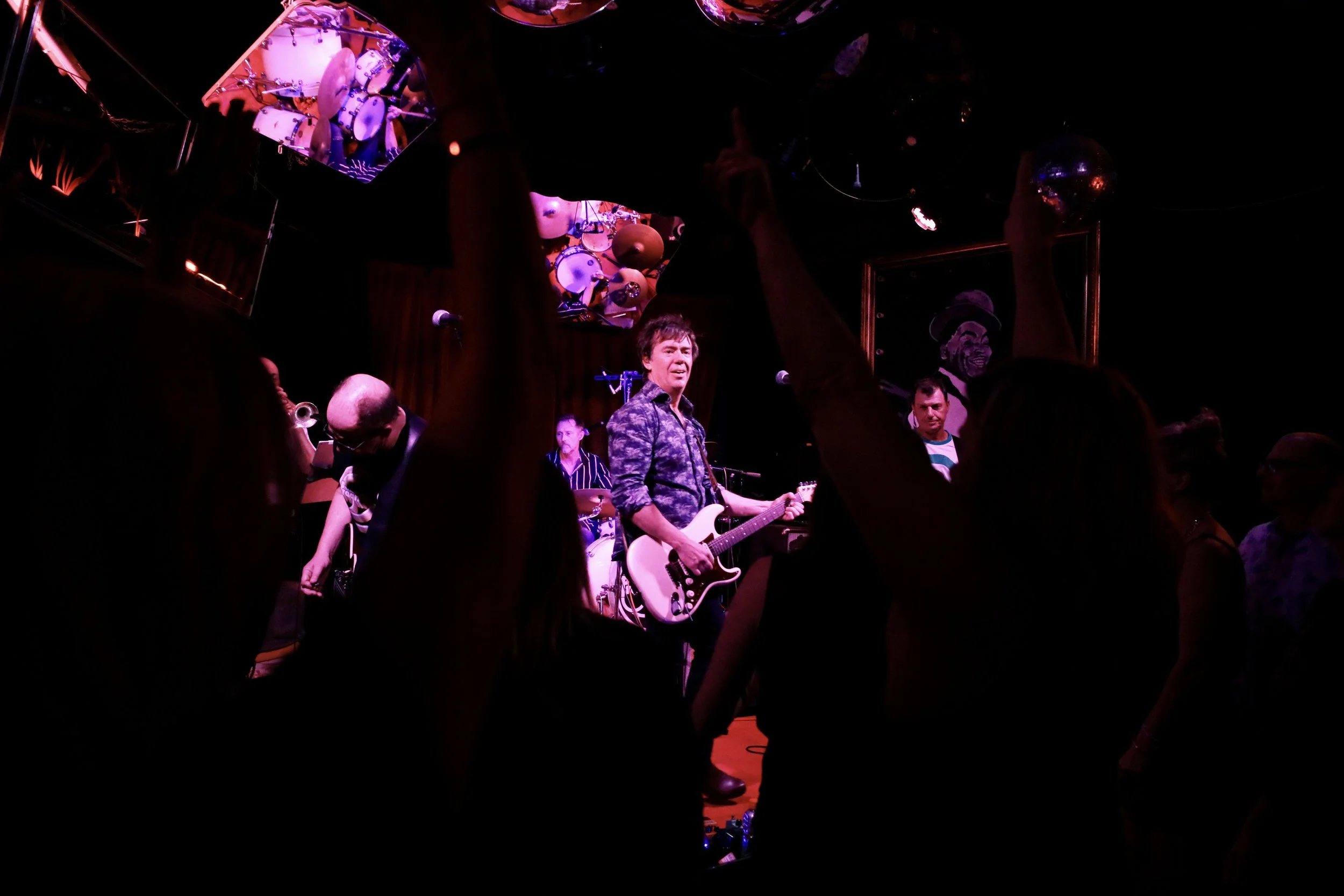 A live music performance in a dimly lit venue with a band on stage, featuring a guitarist in the center, and audience members dancing or enjoying the show.