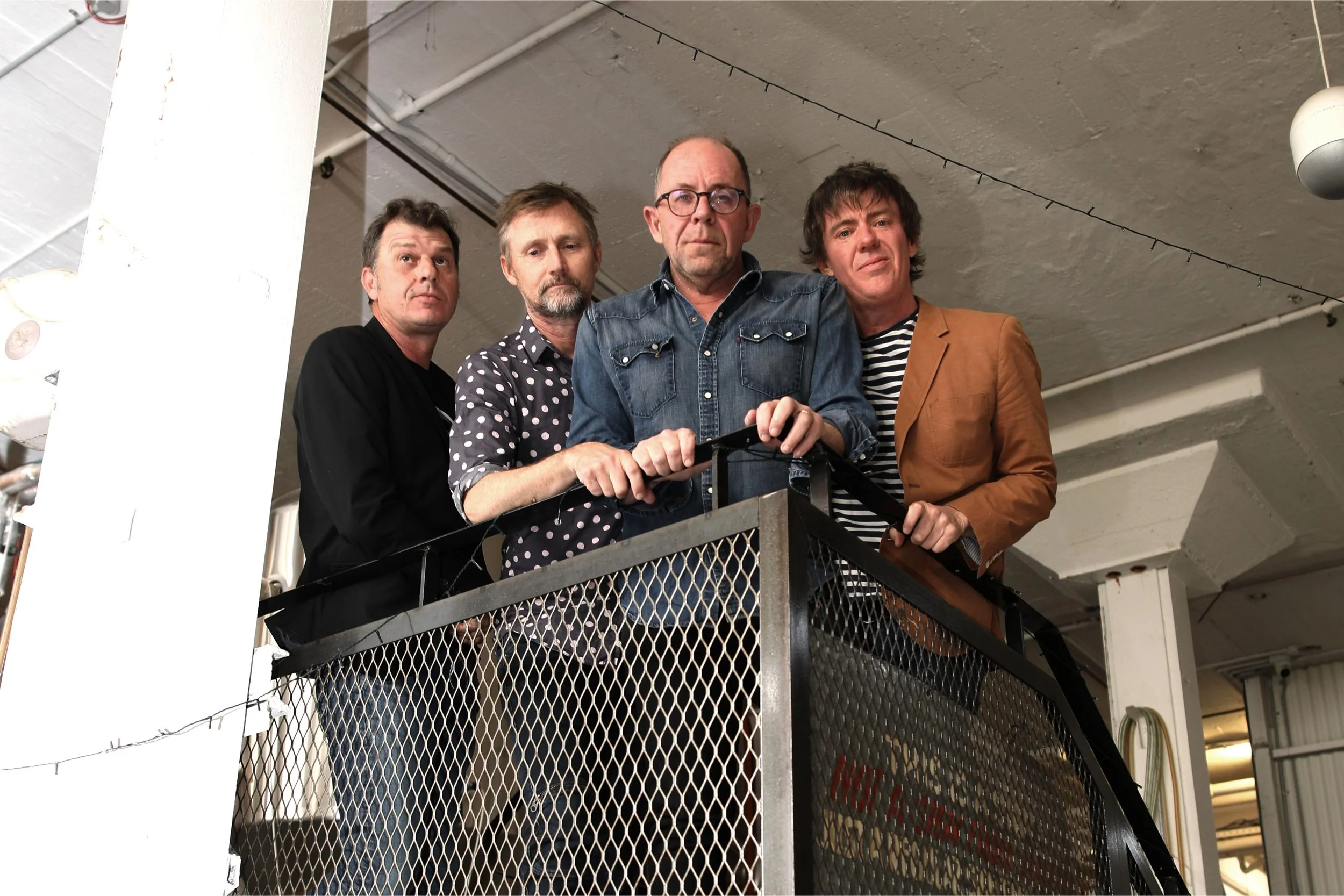 Four men standing side by side on a metal balcony in an industrial-style room.