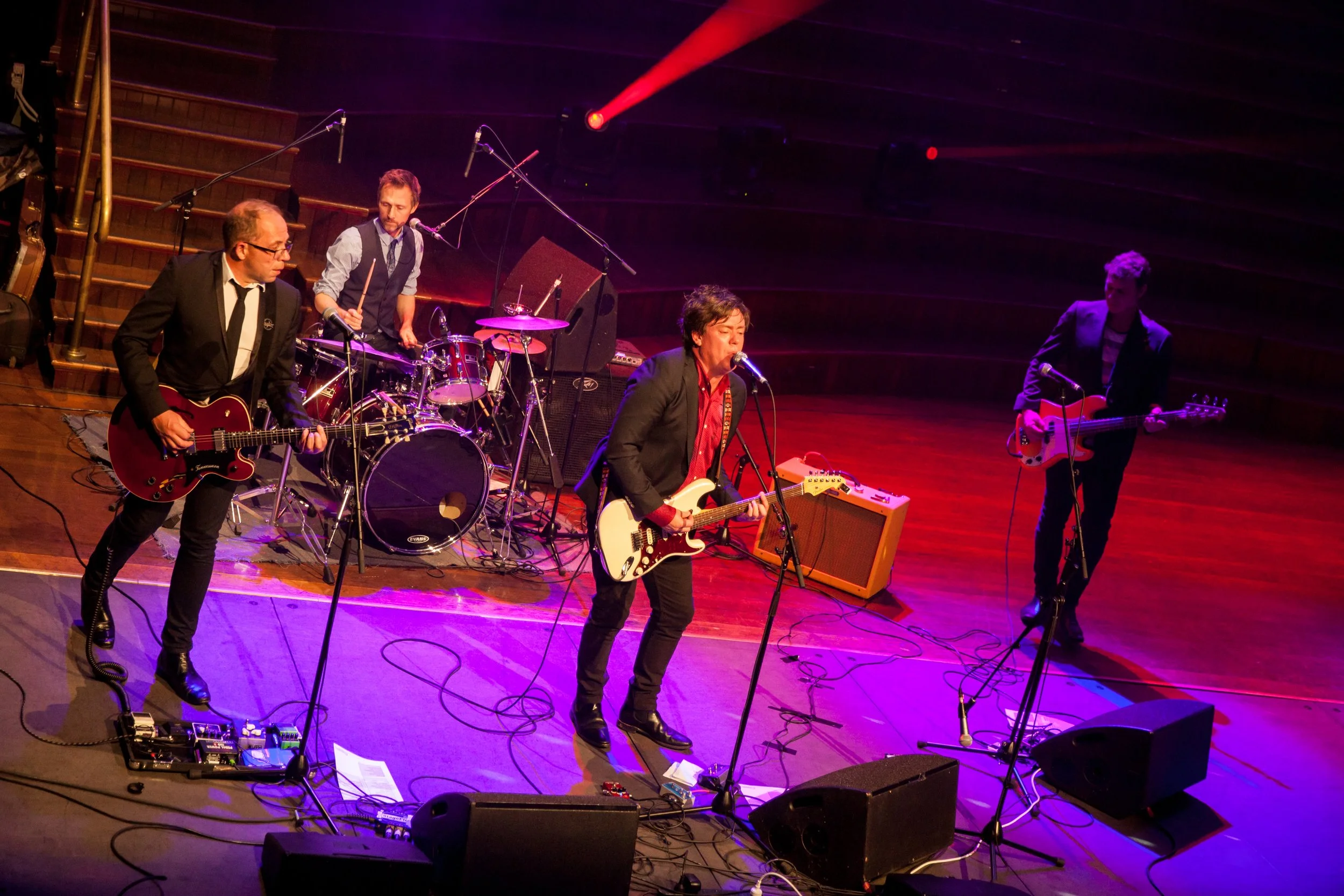 A band performing live on stage with four members playing guitar, drums, and bass. The stage is lit with colorful lights and musical equipment is visible.