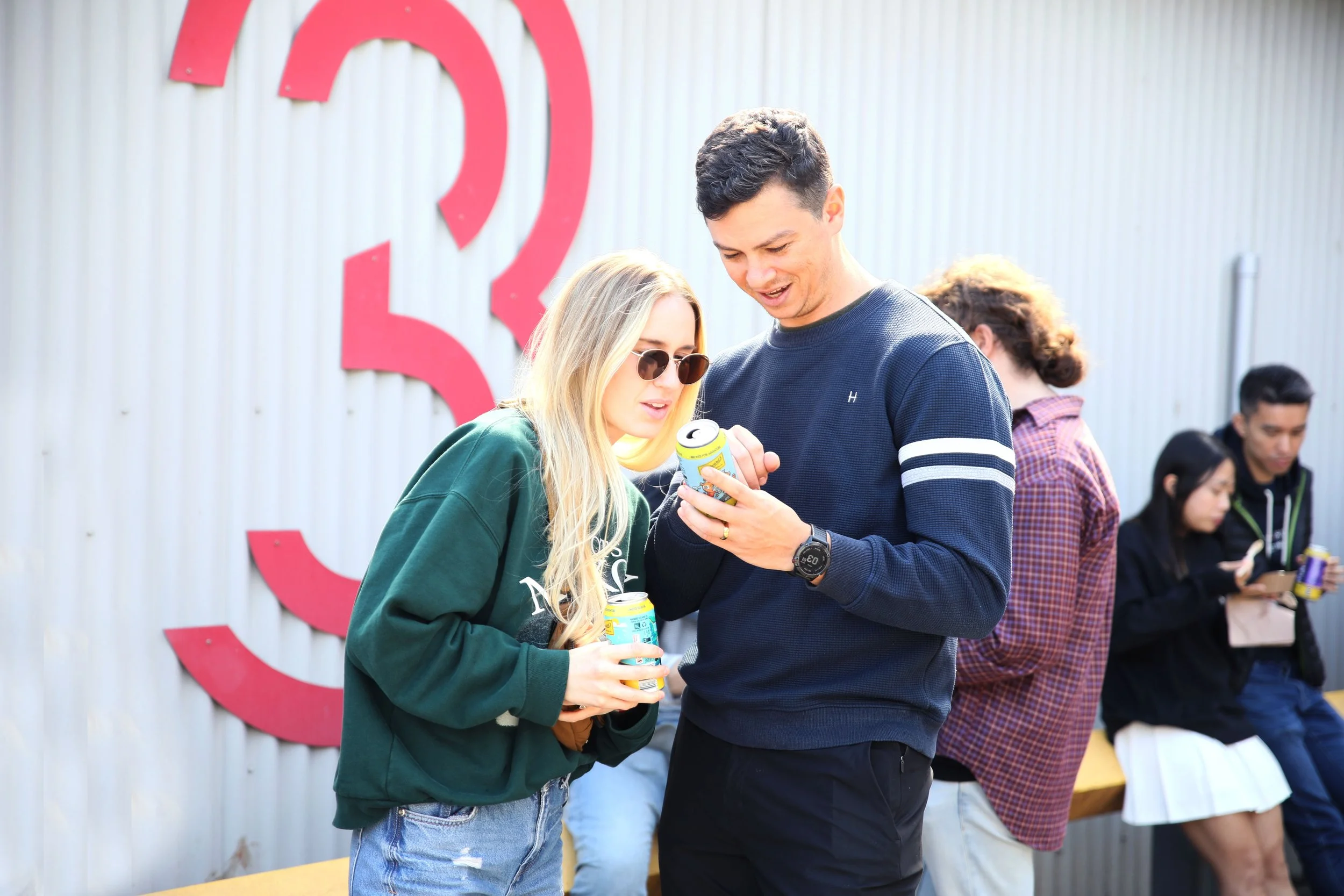 A group of young people standing outdoors, two of them looking at a soda can and engaging in conversation, near a wall with a large red logo.