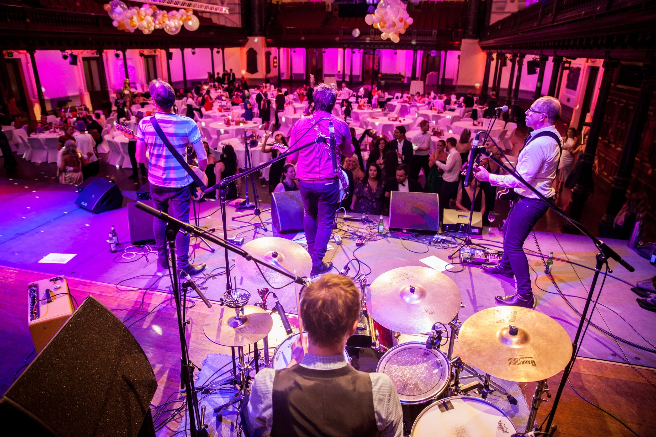 Band performing on stage at an indoor event with purple lighting, audience dancing and socializing in a decorated ballroom.