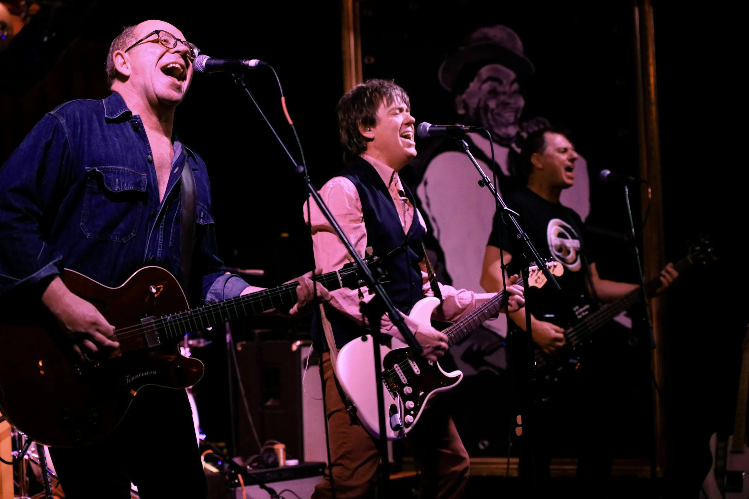 Four musicians performing on stage, singing into microphones. They are playing guitars and appear energetic, with a large portrait of a smiling person wearing a hat in the background.
