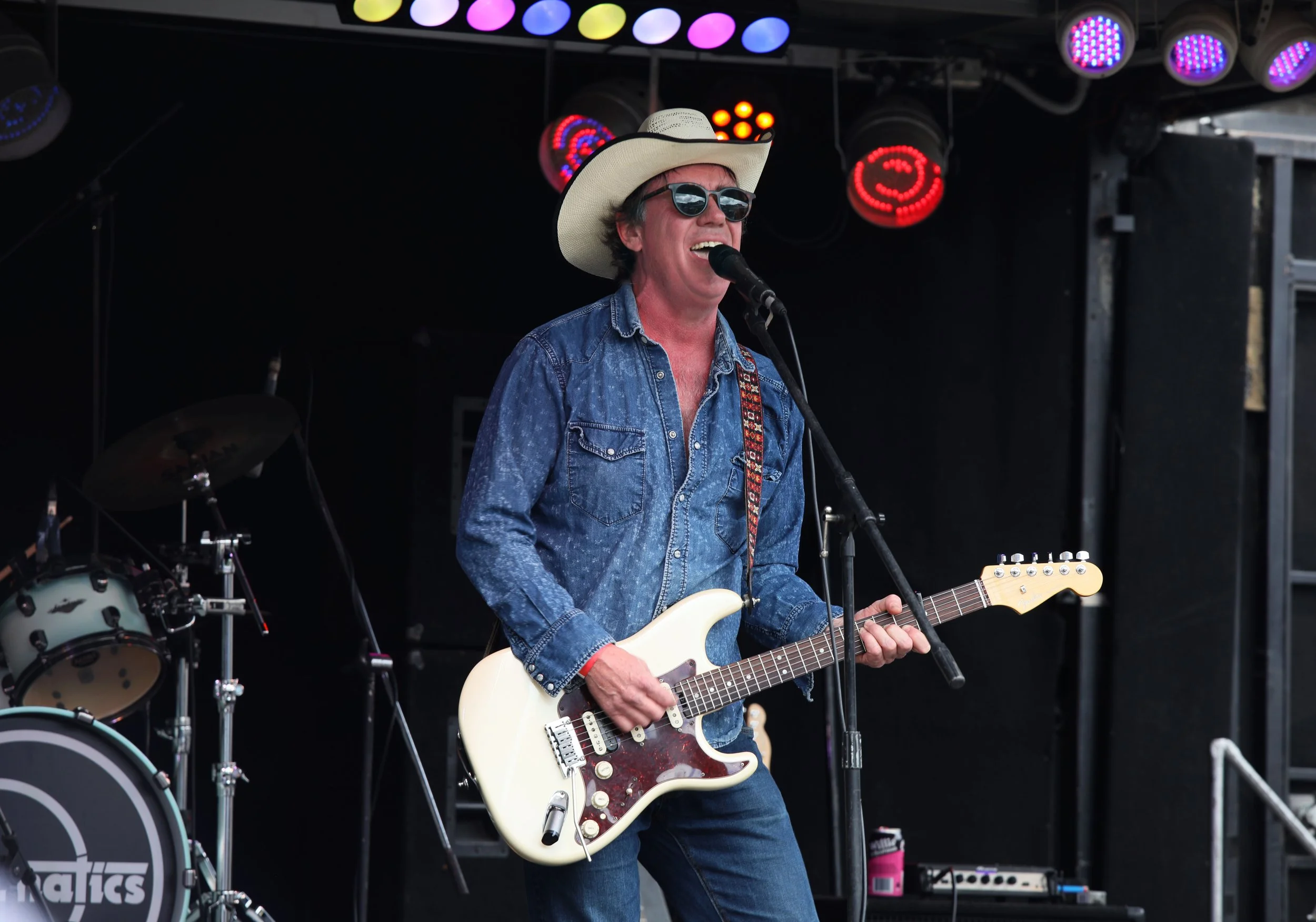 A man wearing sunglasses, a cowboy hat, and a denim shirt playing an electric guitar and singing into a microphone on stage, with colored stage lights above him and a drum set in the background.