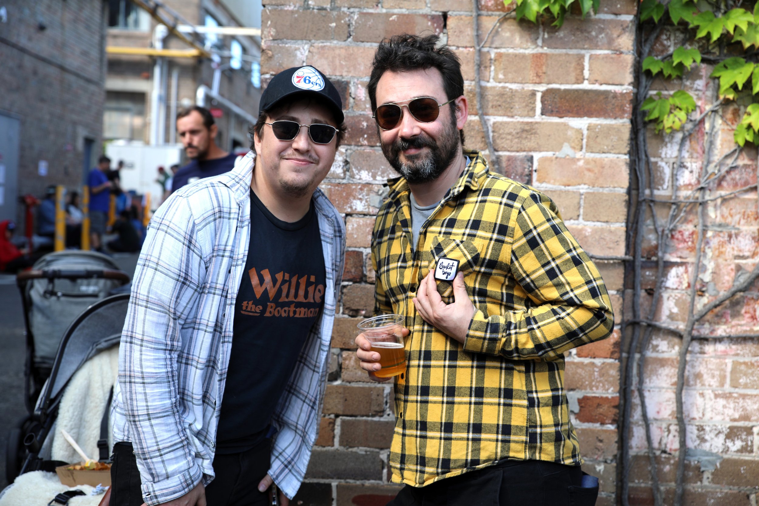 Two men wearing sunglasses standing close together, smiling, with a brick wall and green ivy in the background. The man on the left wears a baseball cap and a black "Willie the Boatman" T-shirt, while the man on the right wears a yellow and black pla