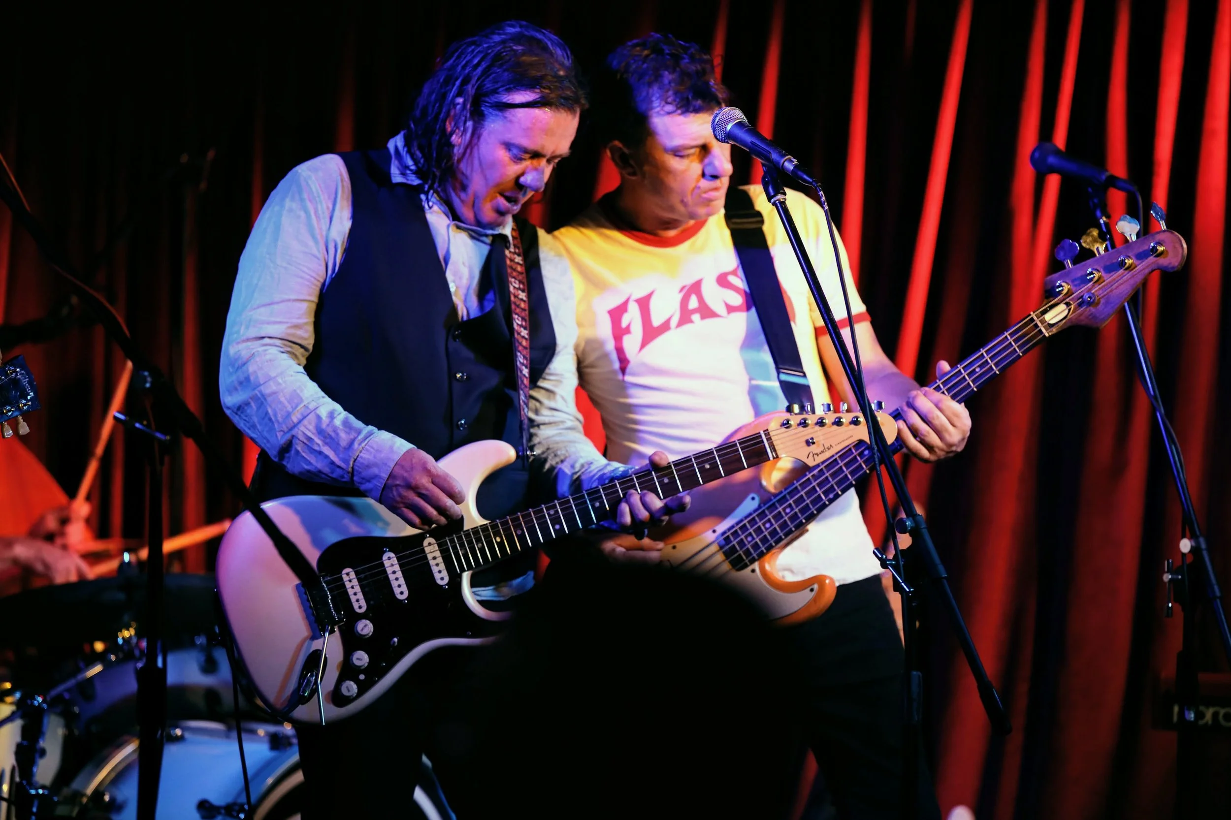Two men playing electric guitars on stage with red curtains behind them.