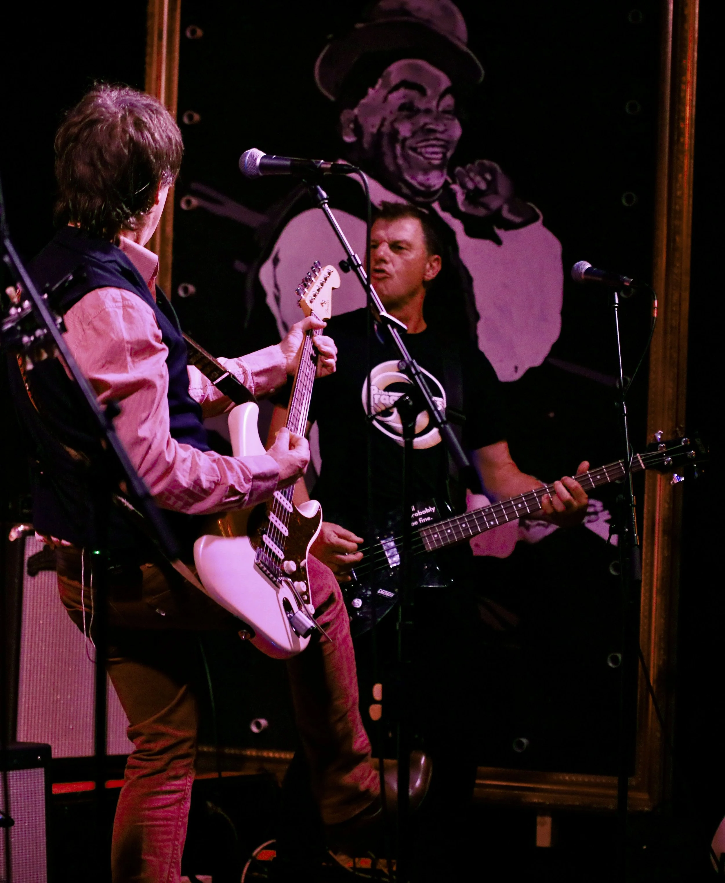 Two musicians playing electric guitars on stage with a large jazz-themed painting in the background.