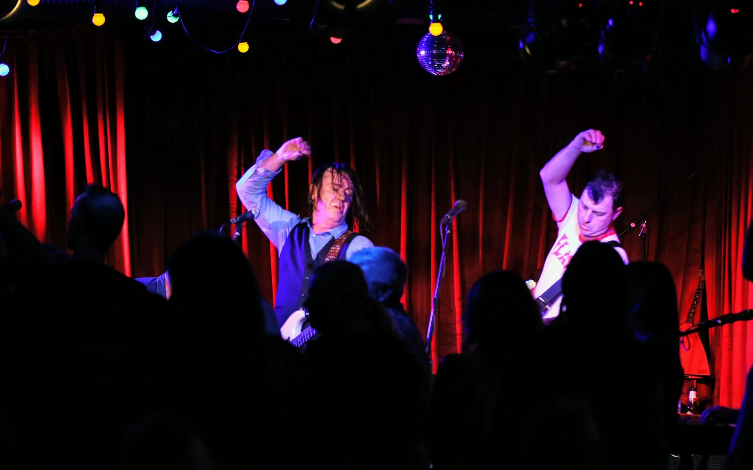 Musicians performing on stage with guitar, dancing, and raising their arms in front of a red curtain with colorful string lights and a disco ball.