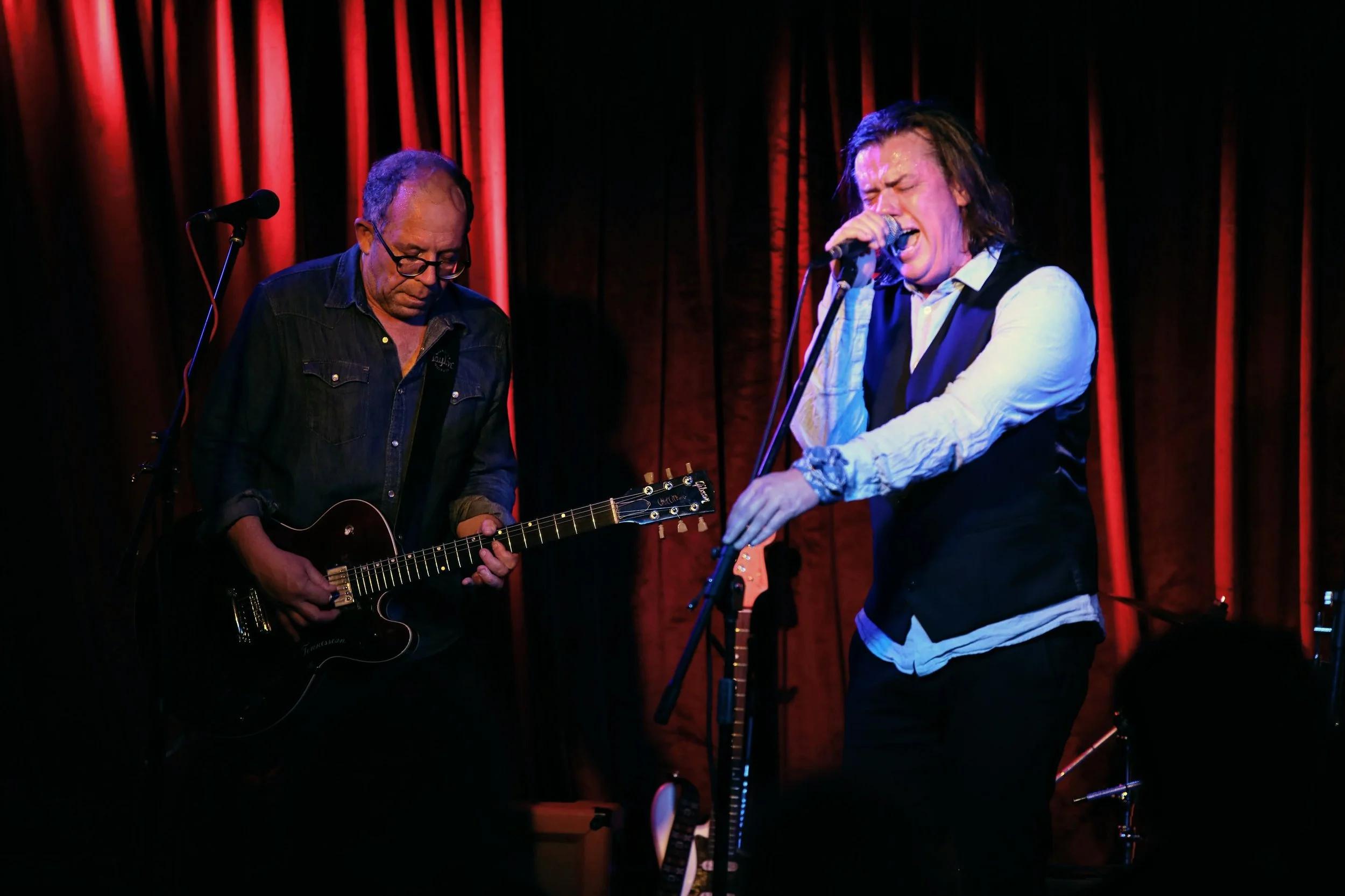 Musicians performing on stage with red curtains, one playing guitar and the other singing passionately into a microphone.
