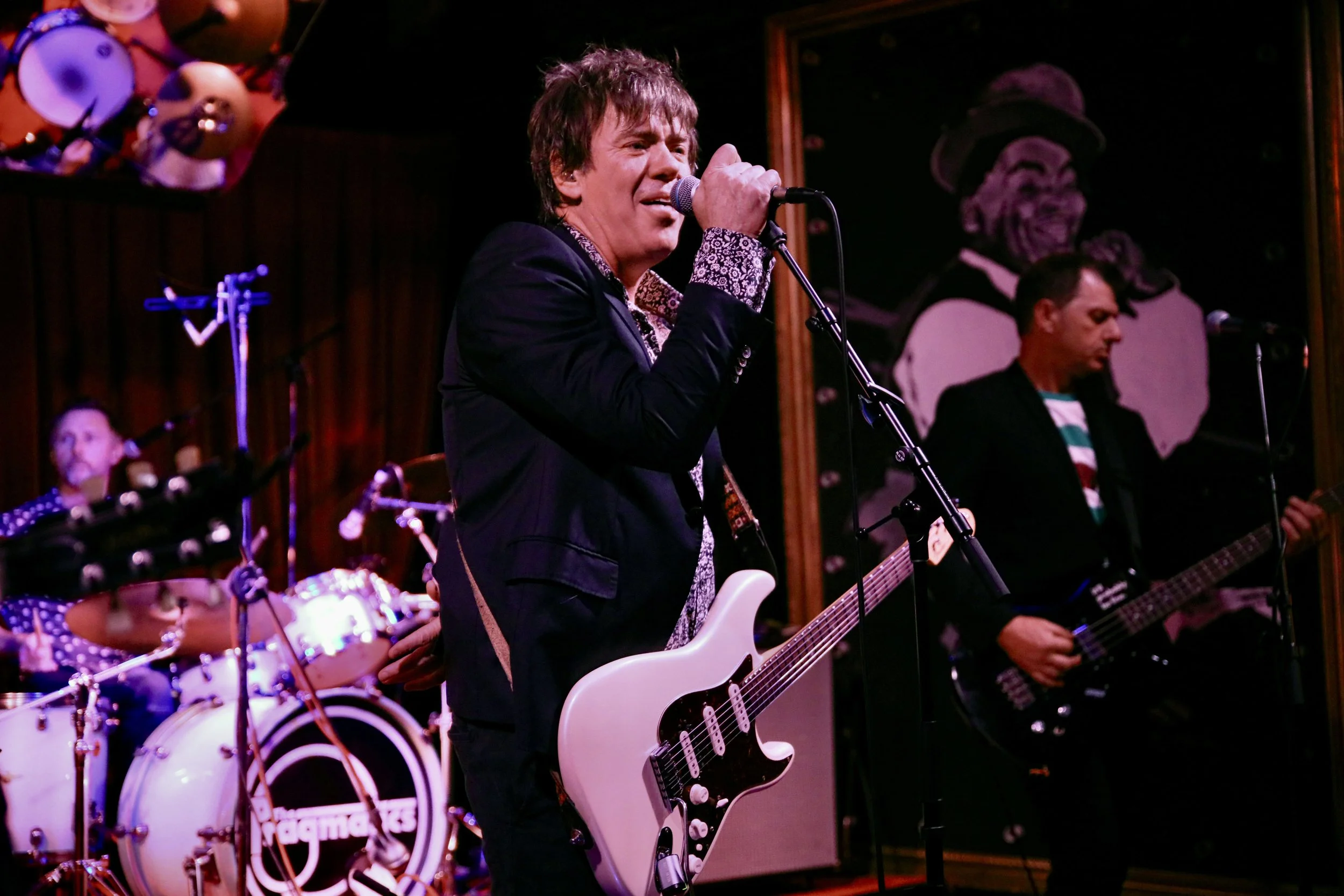 Musicians performing on stage, with a drummer in the background, a singer with a guitar in the center, and a bassist on the right, in a dimly lit venue.