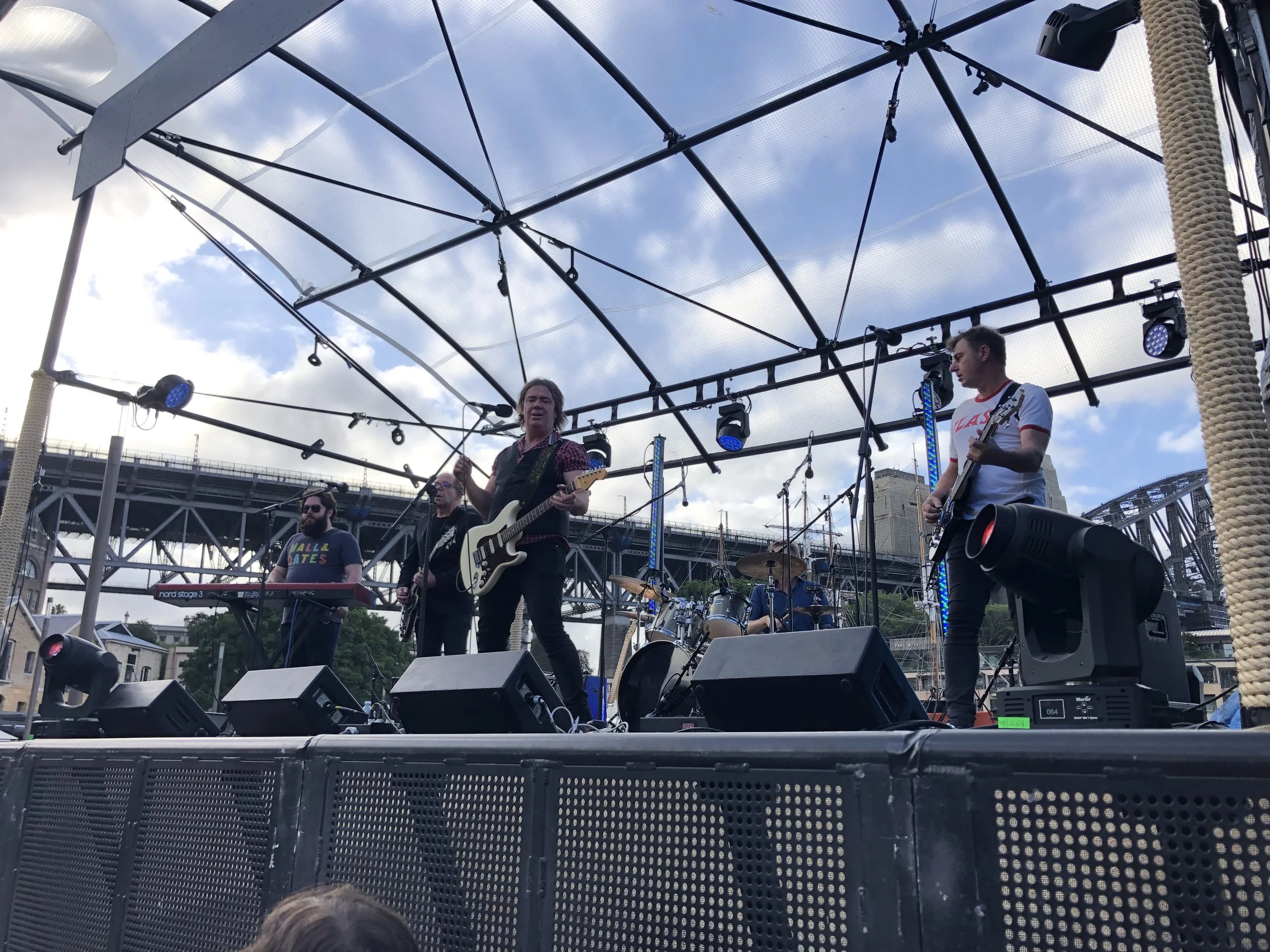 Band performing live on an outdoor stage with a keyboard, guitars, and drums, under a partly cloudy sky.