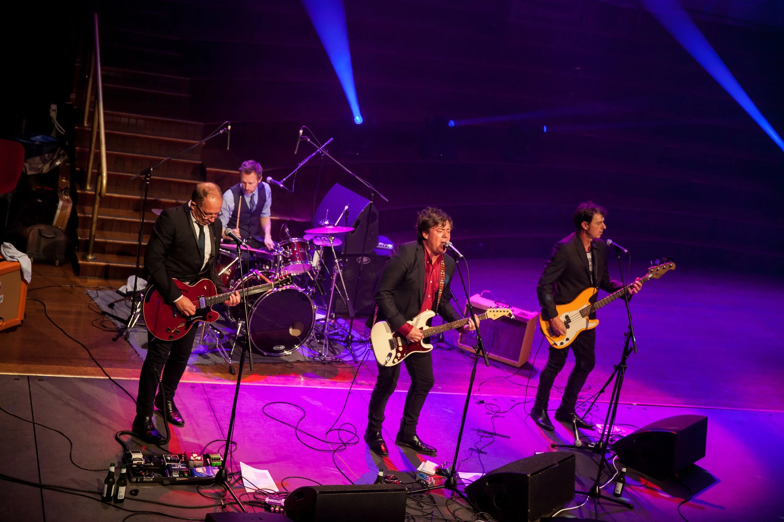 A band of four musicians performing on stage with purple and blue lighting.