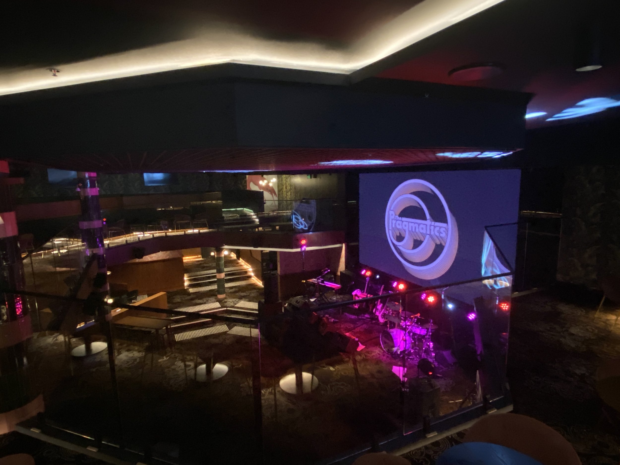 Empty concert venue with a stage set up with musical instruments, colorful stage lights, and a large screen displaying the logo 'The Pragmatics.' The venue has multiple levels and curving architectural features.