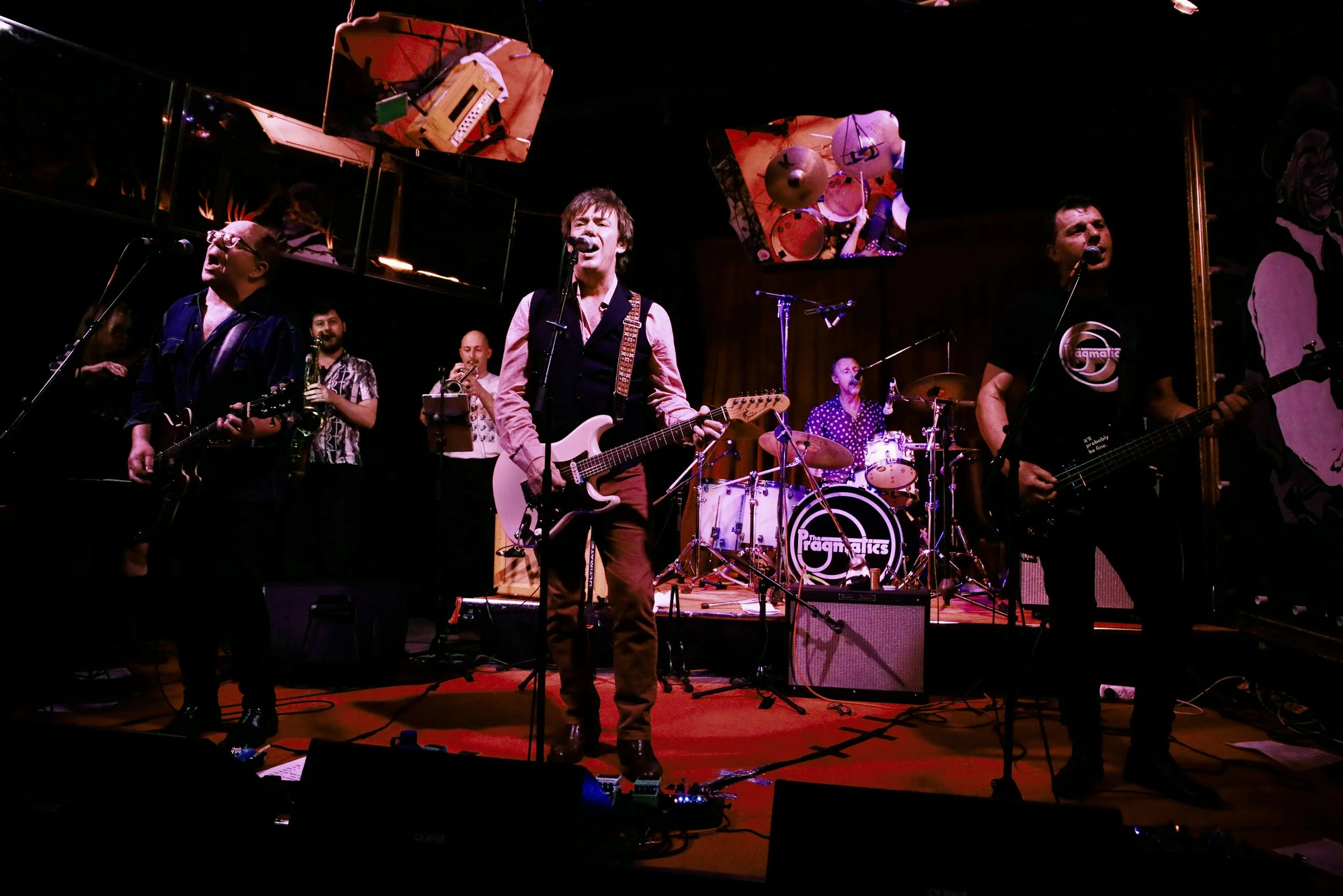 A band performing live on stage with five members: two guitarists, a drummer, a saxophonist, and a bassist. The stage has colorful artwork and mirrors in the background, with the band's name visible on the drum kit, and stage lighting highlighting th