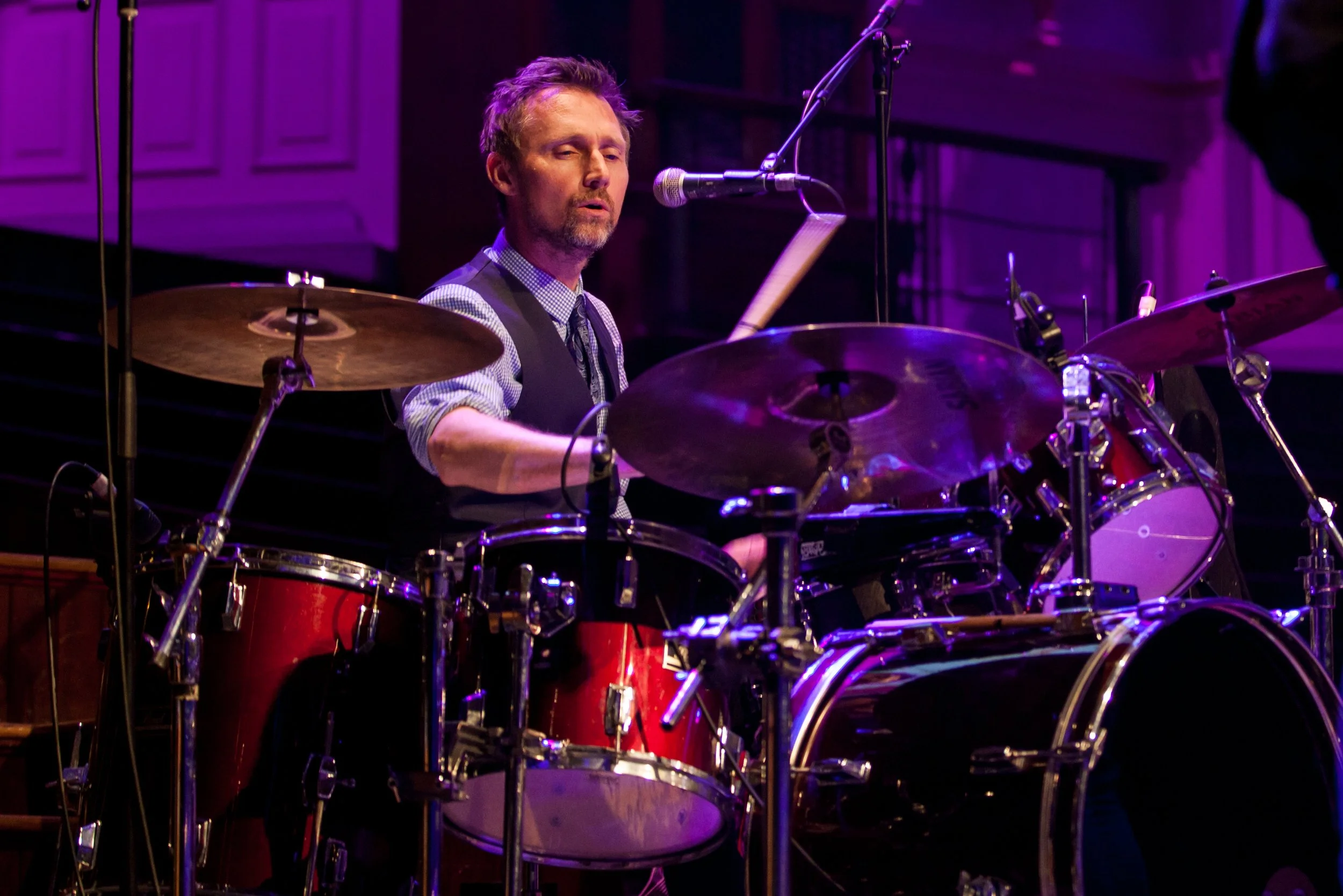 A man playing a drum set on stage, surrounded by drums and cymbals, with purple stage lighting.