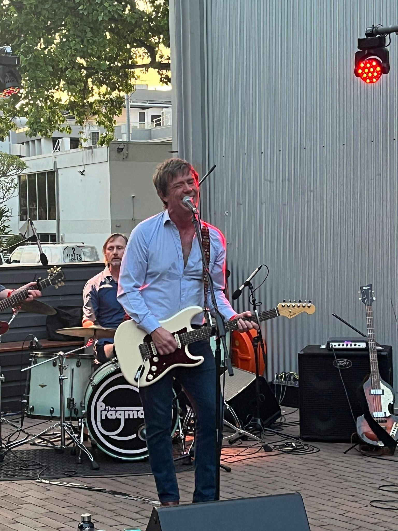 A band performing outdoors, with the lead singer playing an electric guitar and singing into a microphone. The drummer is visible in the background, and the drum set has a logo that reads 'The Pragmas'. There are other guitars and equipment on stage, with a red stage light overhead.