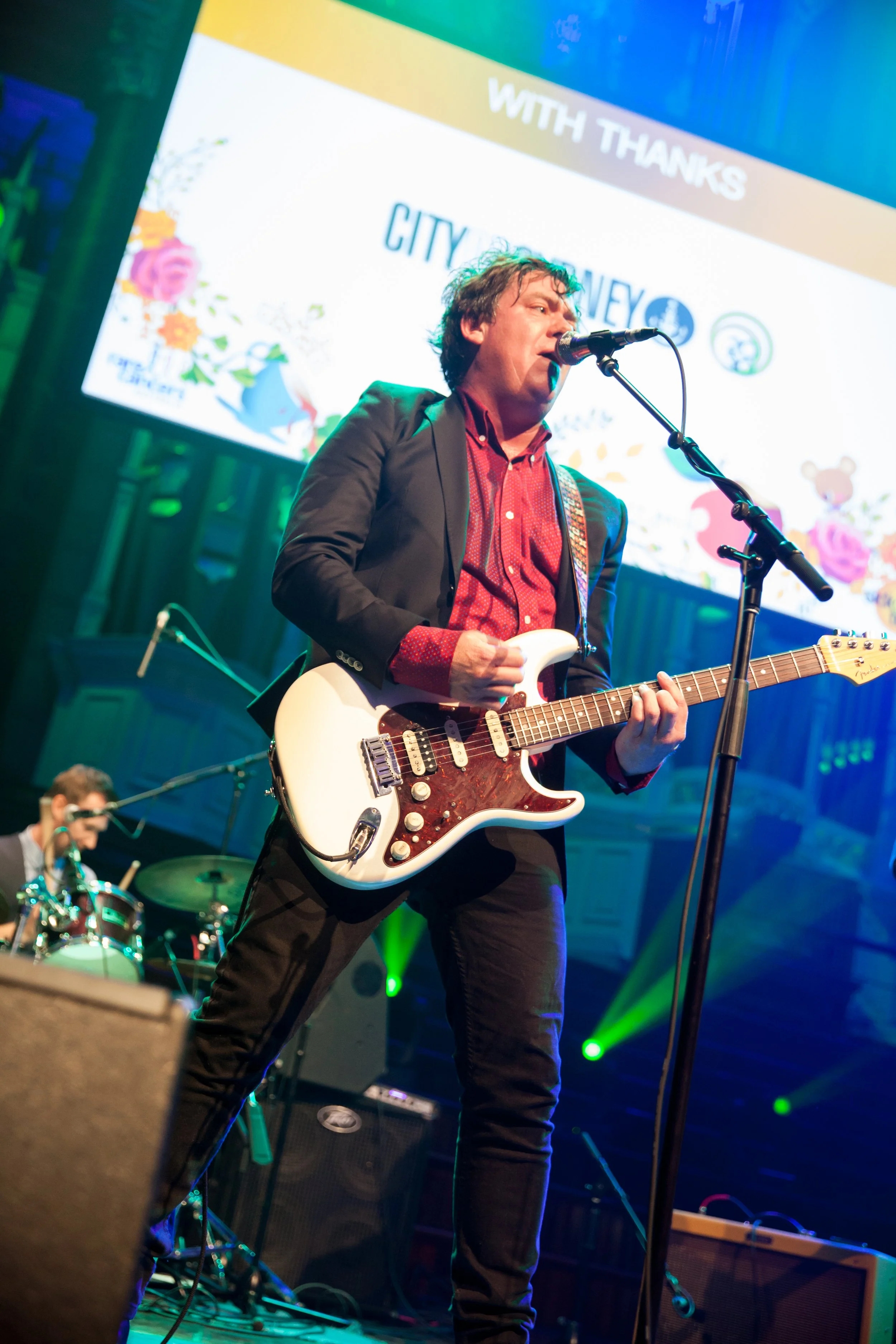 A male musician with dark curly hair playing an electric guitar on stage, singing into a microphone, with a drummer in the background and a large screen displaying a colorful graphic in the background.