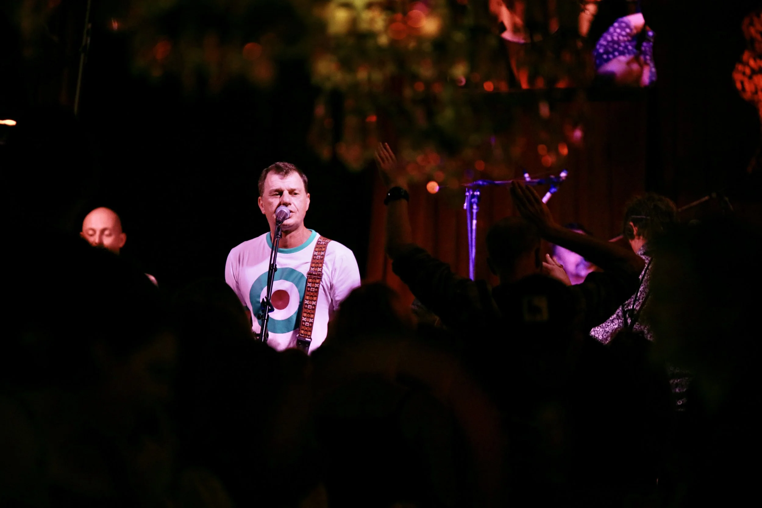 A man singing into a microphone on stage, wearing a T-shirt with a colorful concentric circle logo, while audience members raise their hands in front of him. The background is dark with some lights and decorations.