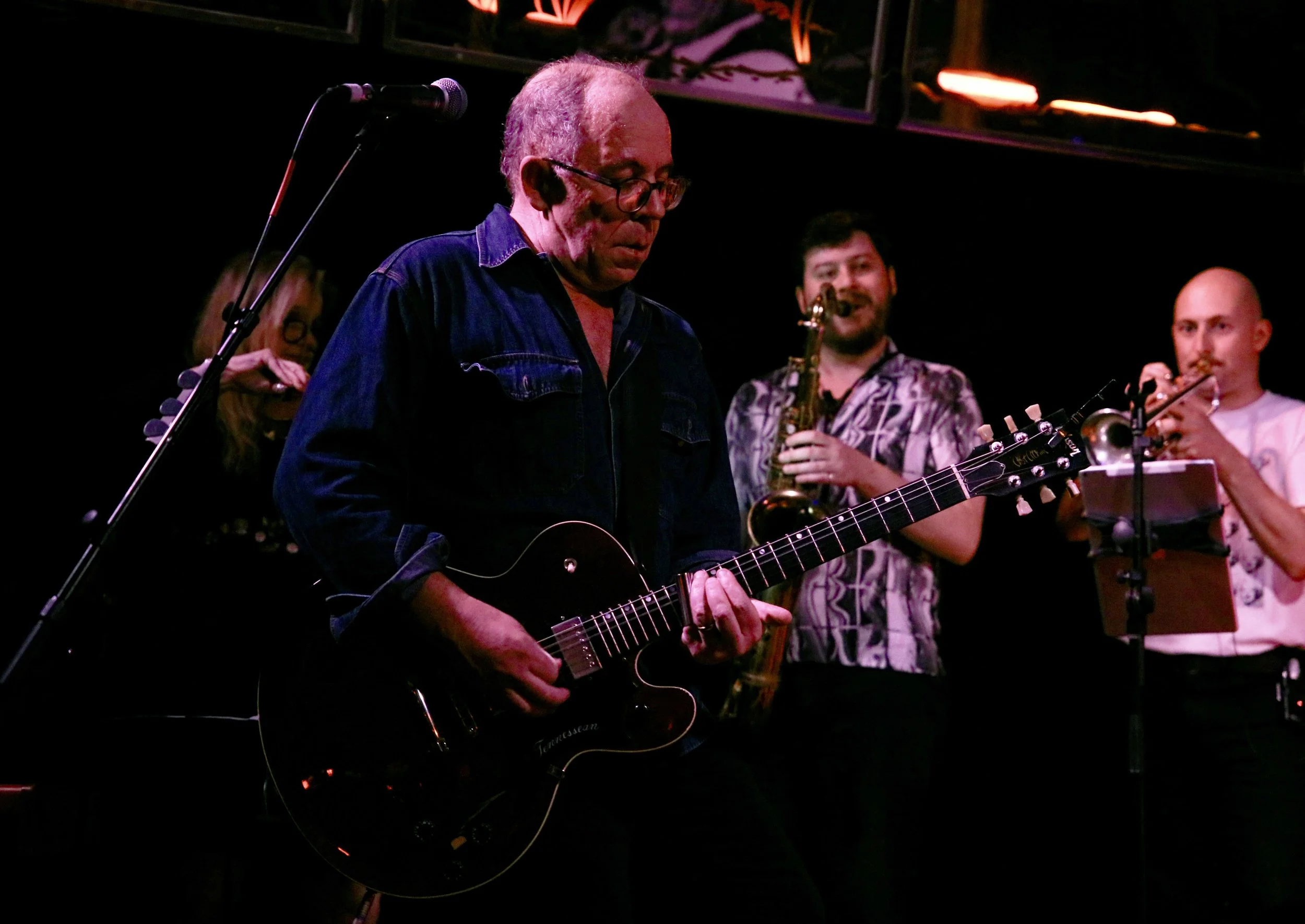 A man playing an electric guitar on stage during a live performance, with other musicians in the background, including a saxophonist and a flutist.