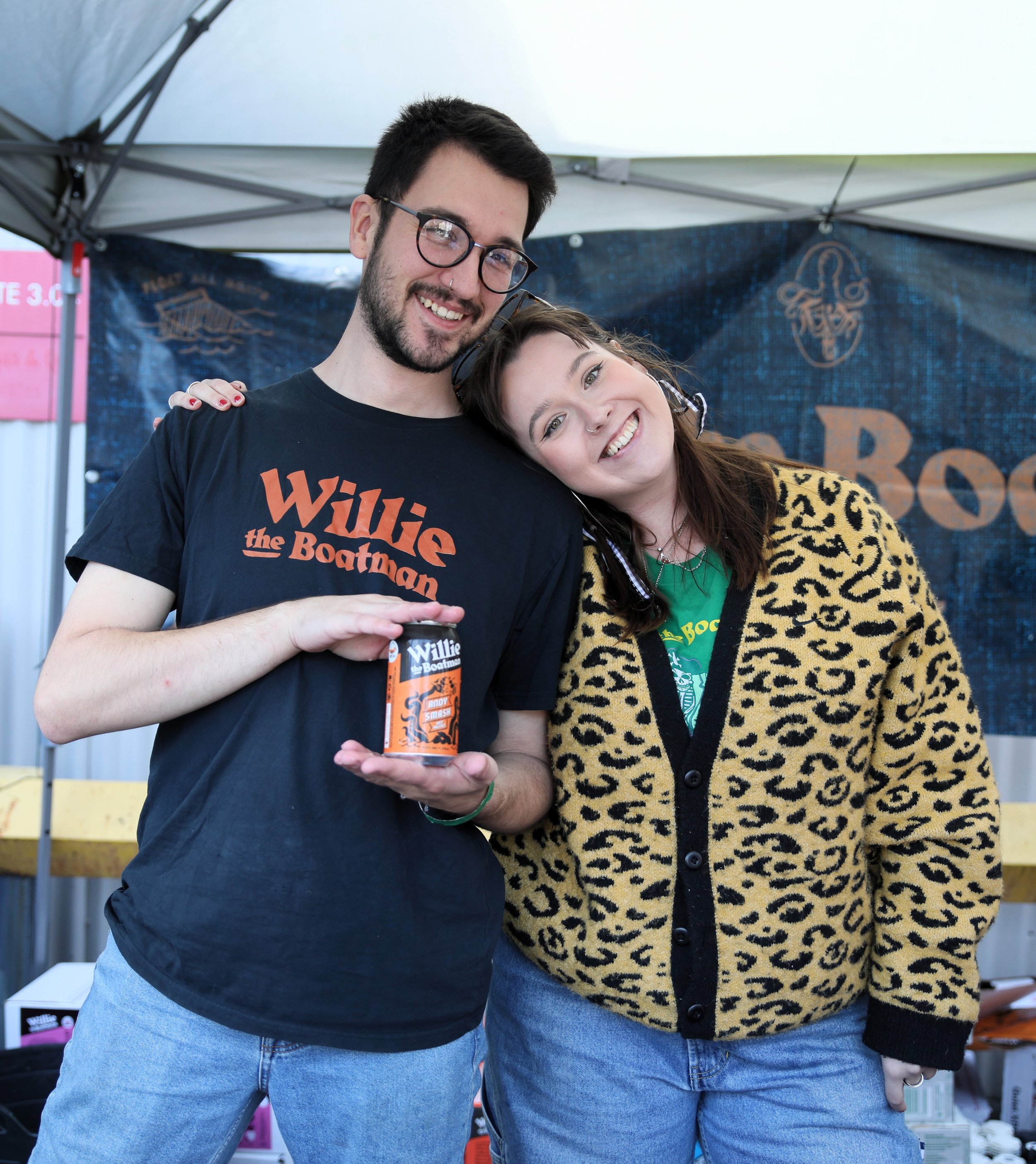 A man and woman smiling at a booth, the man wearing a black t-shirt with orange text 'Willie the Boatman' and holding a can, the woman wearing a yellow leopard print cardigan and a green t-shirt underneath.