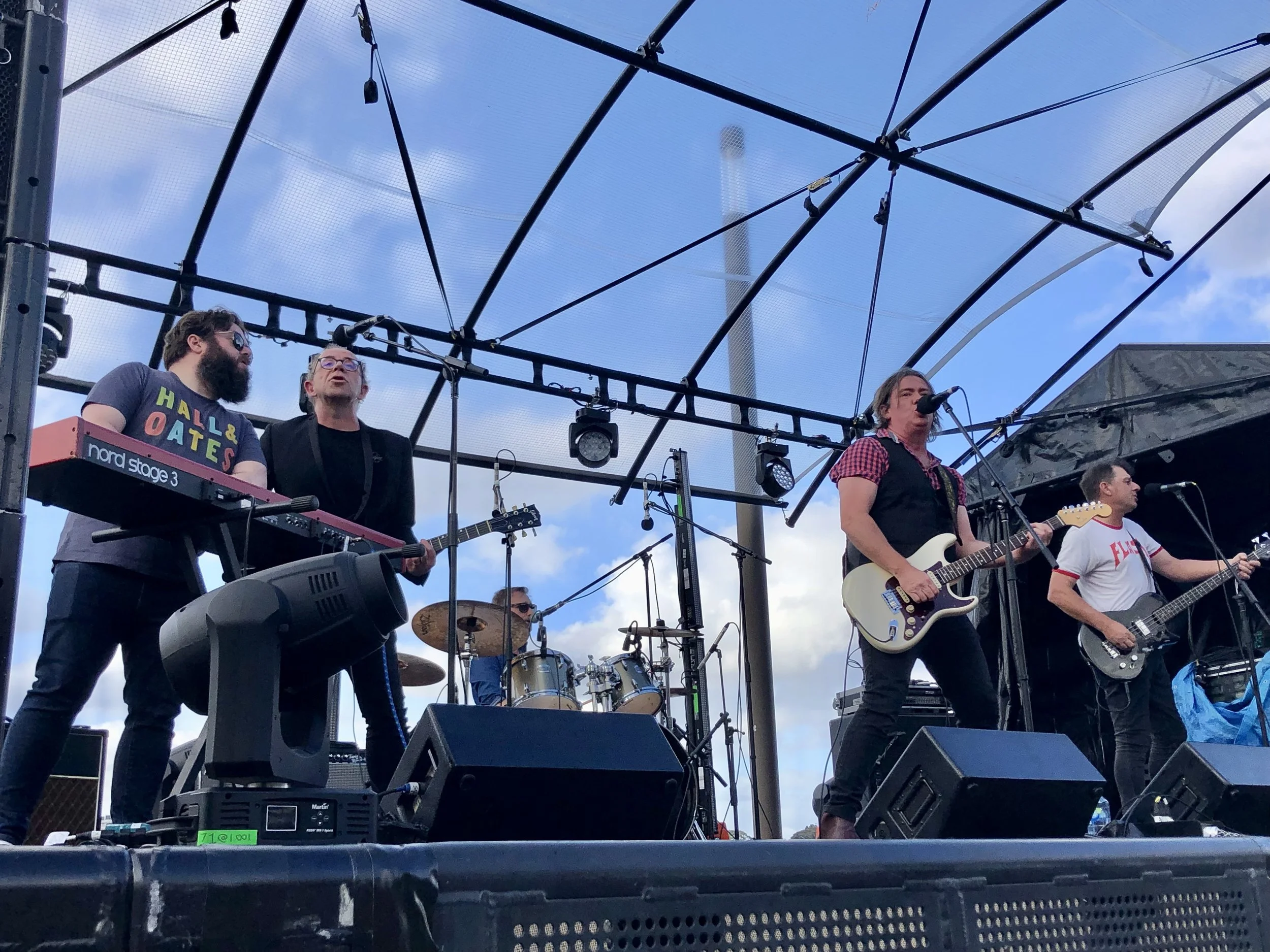 A band performing on an outdoor stage with a blue sky and clouds in the background. The band includes a keyboardist, a drummer, and two guitarists, one of whom is singing into a microphone.