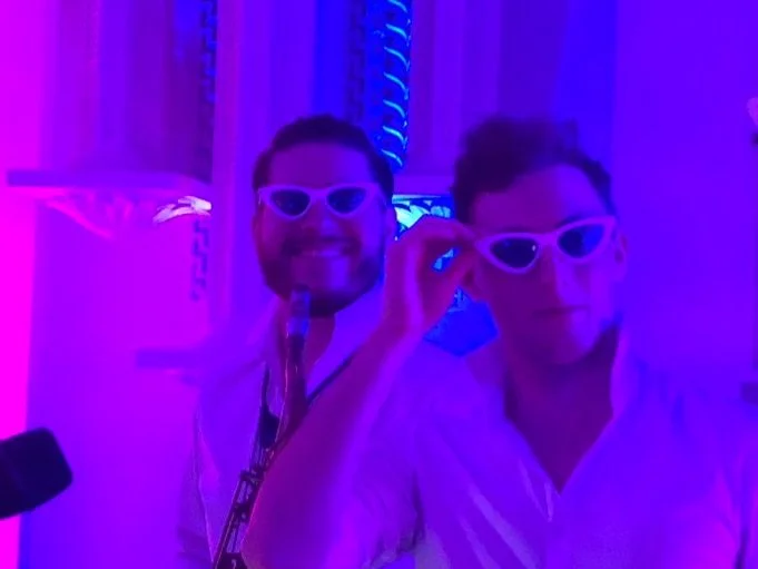 Two men in white shirts wearing white sunglasses, one smiling and one adjusting his glasses, in a purple-lit environment.