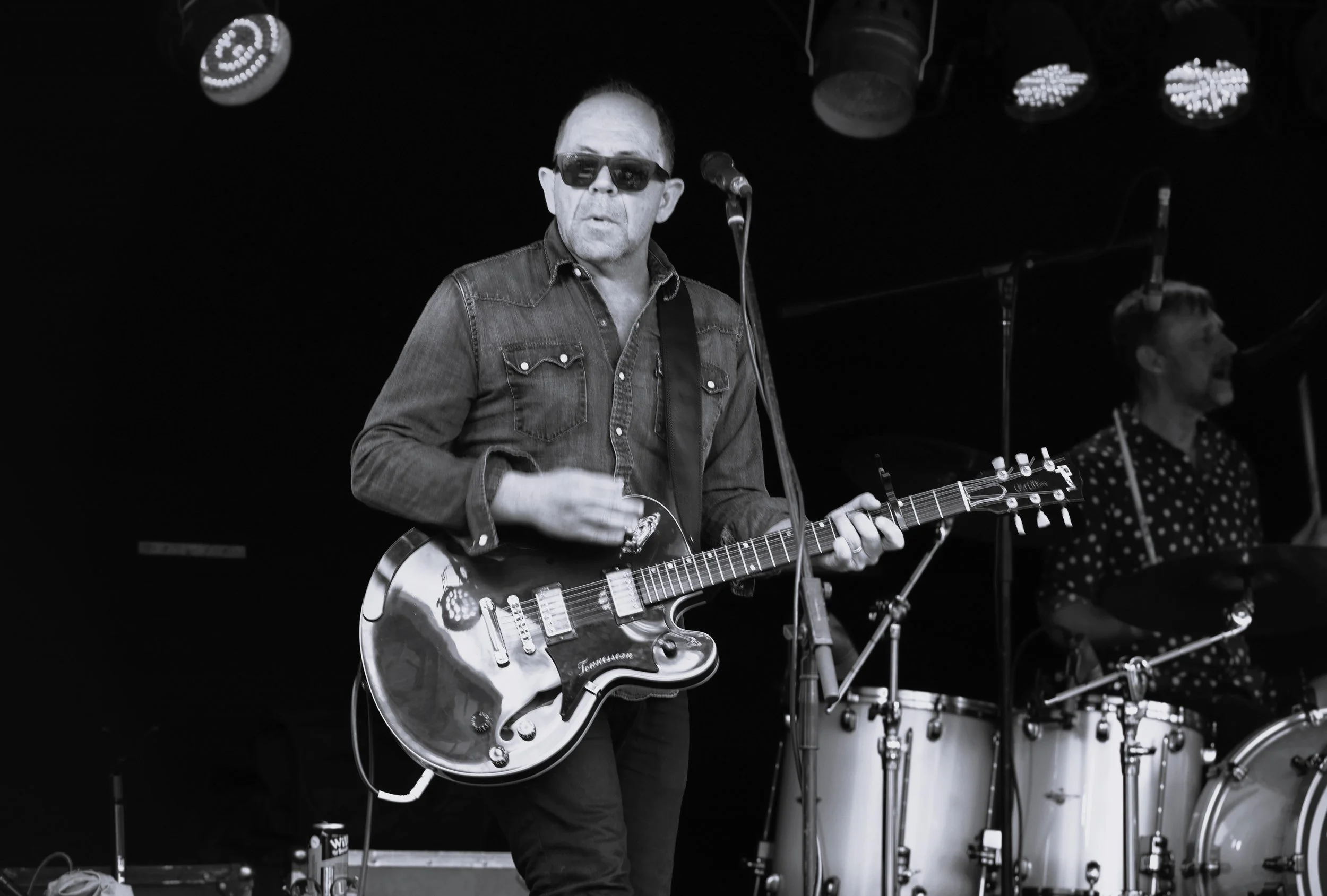 Black-and-white photo of a man playing an electric guitar on stage. He is wearing sunglasses, a denim shirt, and is singing into a microphone. In the background, another musician is playing drums.