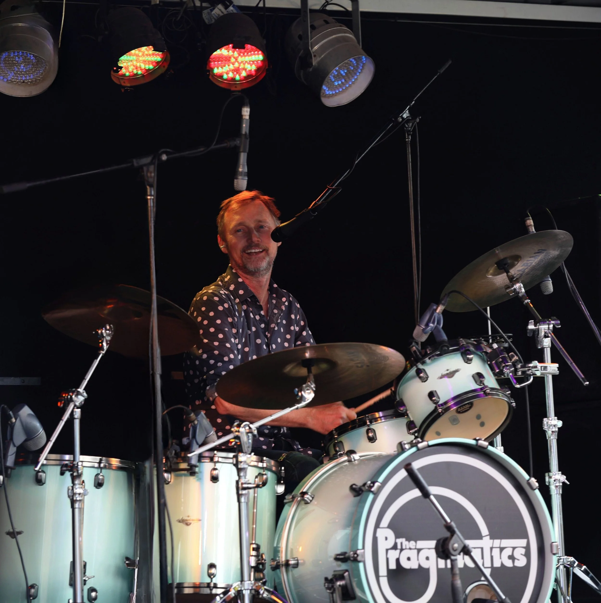 A smiling man playing the drums on stage, wearing a black shirt with white polka dots, underneath colorful stage lights.
