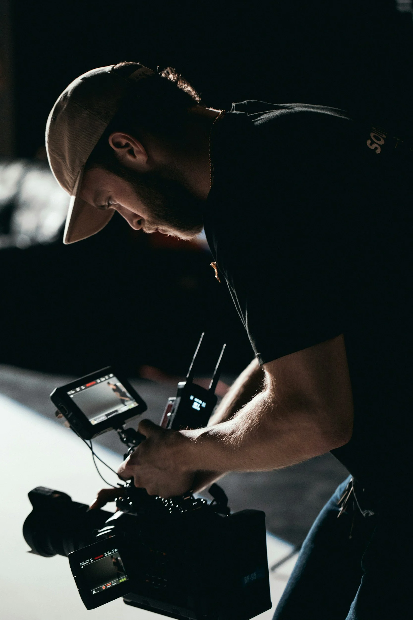 A man wearing a beige hat is operating professional video camera equipment in a dimly lit environment.