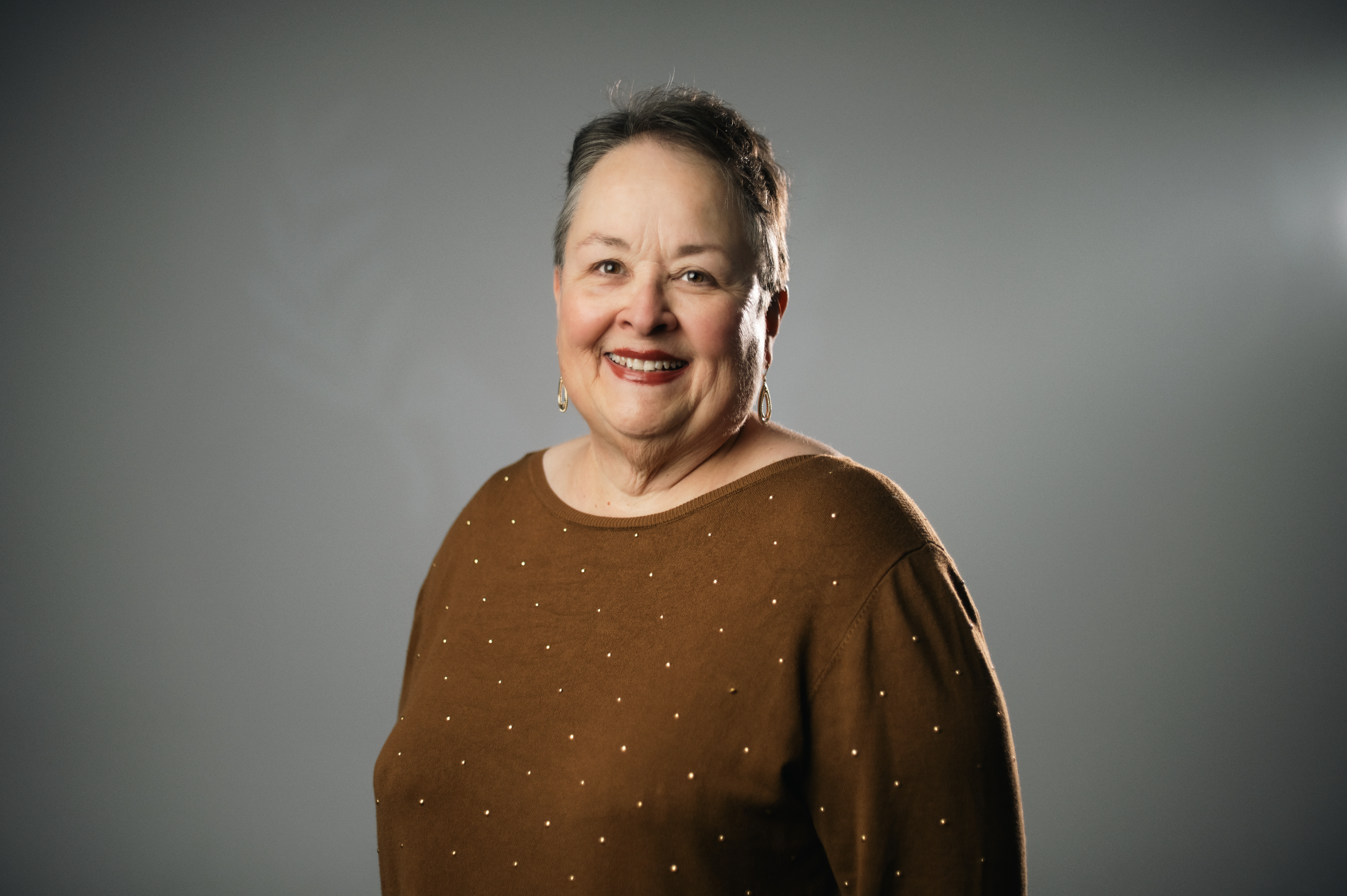 A woman with short gray hair, glasses, and earrings, smiling broadly, wearing a black top with small gold embellishments against a beige background.