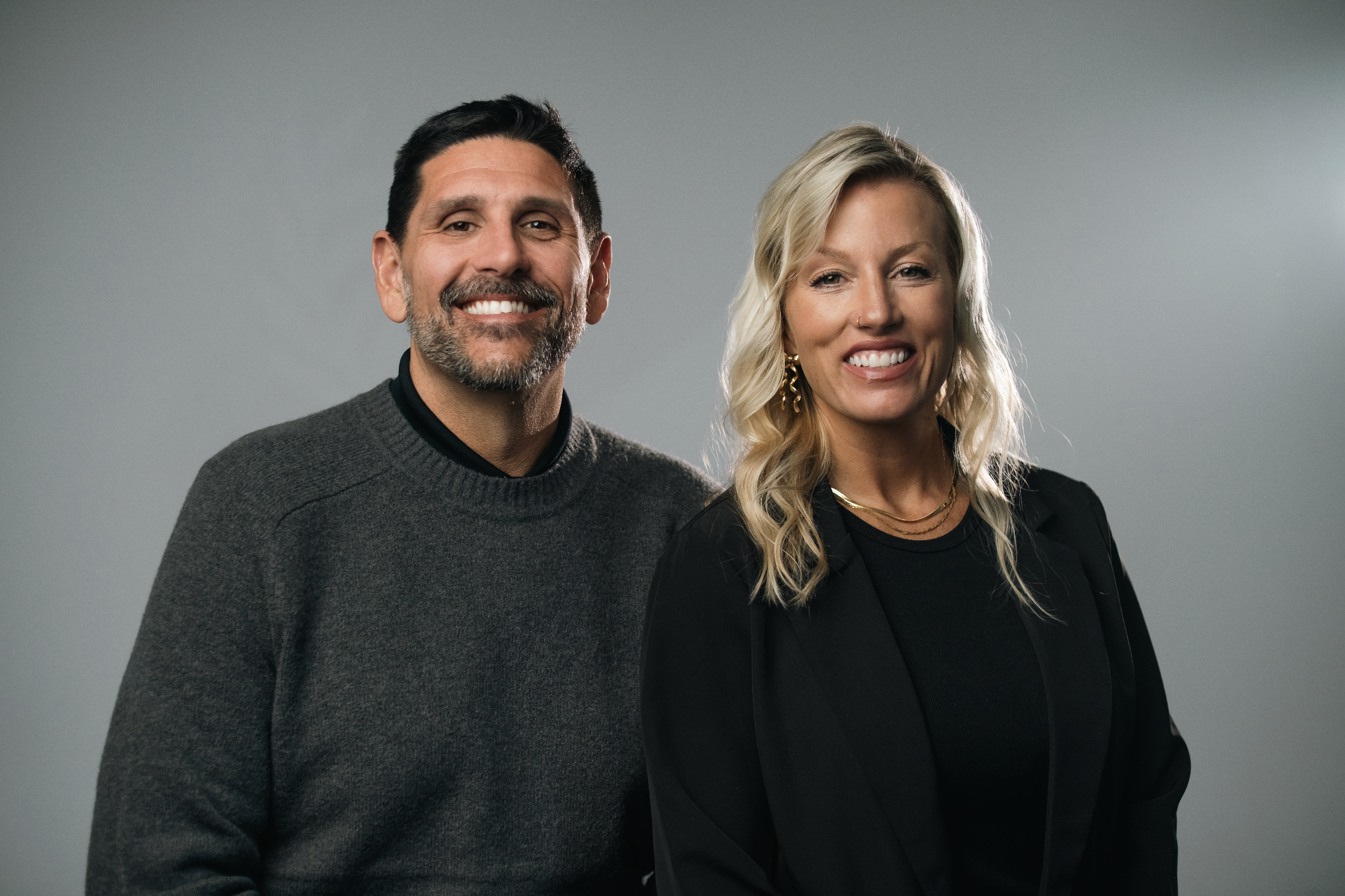A man and woman standing close together, smiling, against a plain beige background.