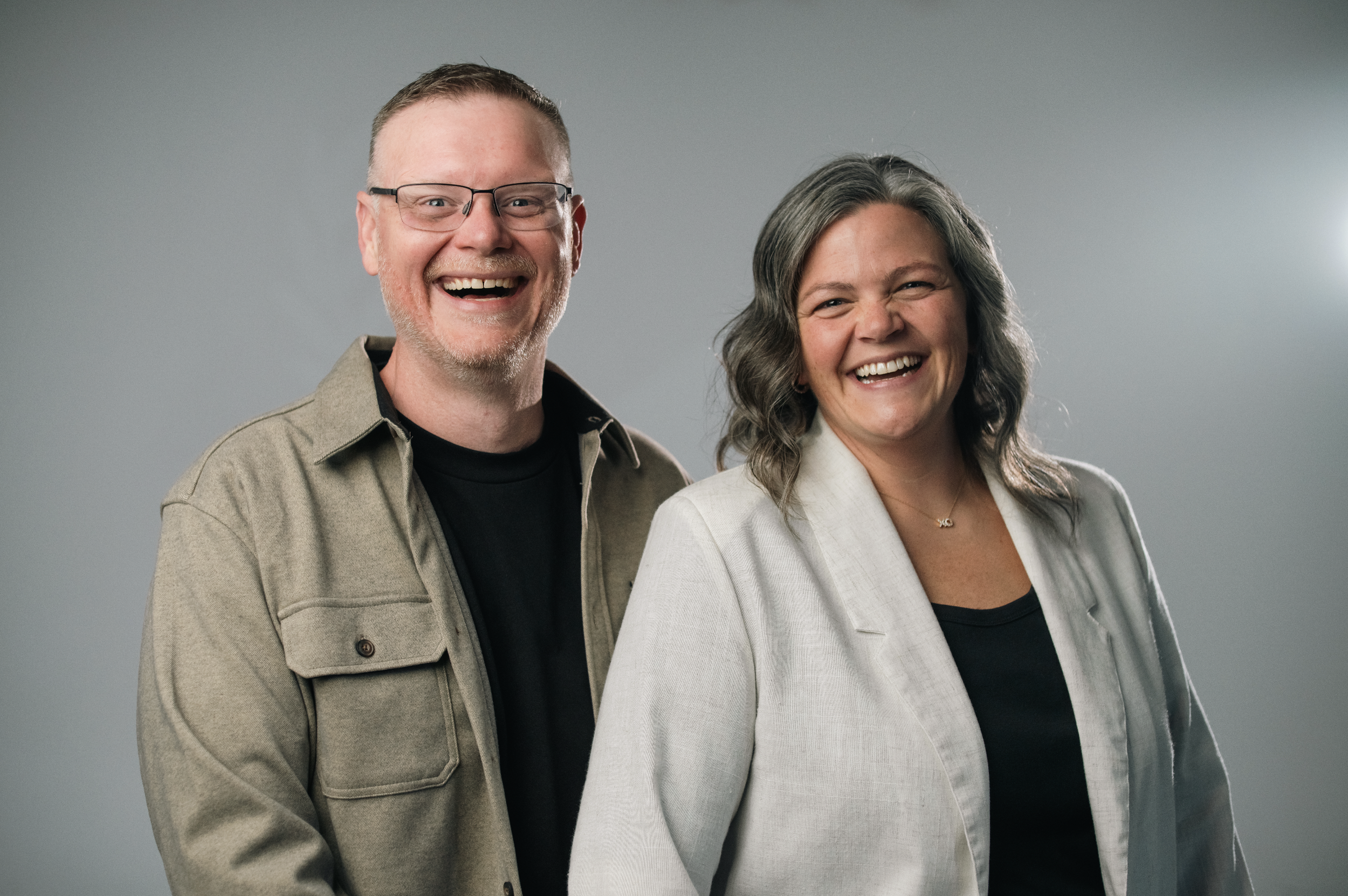 A smiling man wearing glasses and a brown zip-up sweater standing next to a smiling woman with wavy gray hair wearing a white blouse, against a plain beige background.
